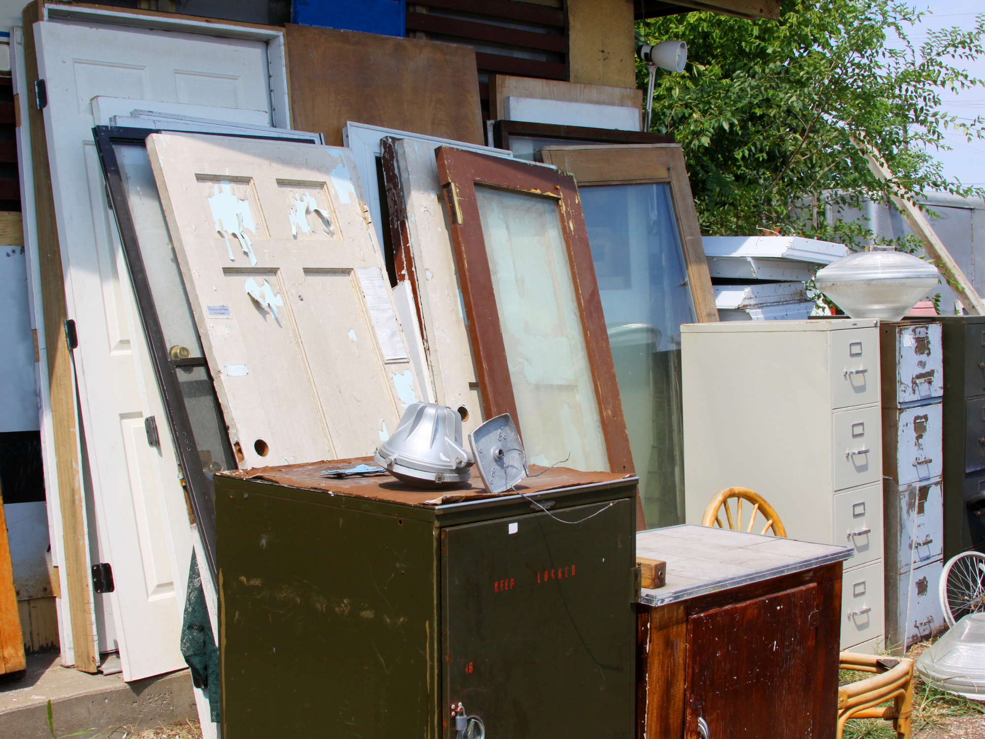 A pile of discarded doors, windows, and old furniture stacked outdoors next to a building, with some green trees in the background.