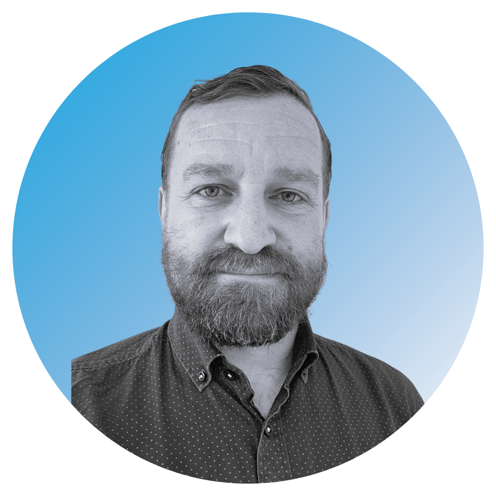 Black and white portrait of a man with a beard and short hair, wearing a buttoned-up, collared shirt with small polka dots, set against a blue circular background.