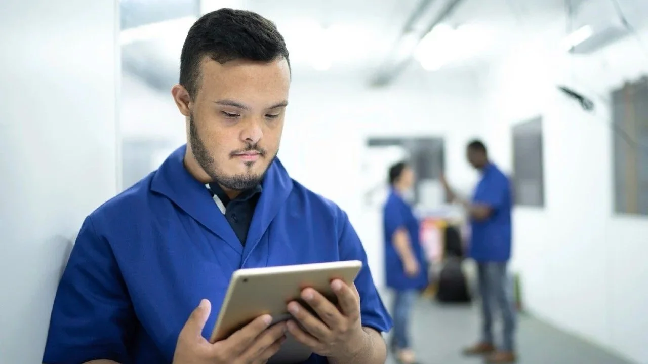 A young man in blue scrubs looking at a tablet in a hospital or medical setting with two other medical staff blurred in the background.