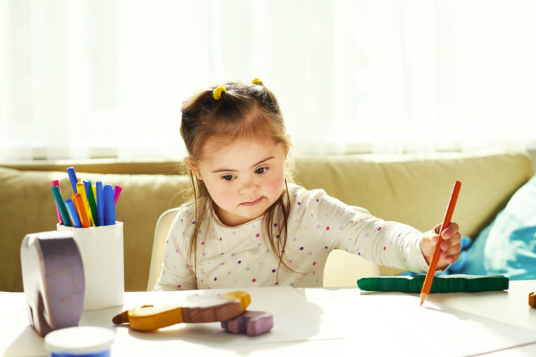 Young girl reaching for an orange crayon at a table with art supplies and erasers in a well-lit room.