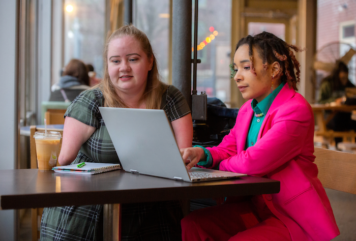 Two women sitting at a table in a cafe, working on a laptop together, with a notebook and iced coffee on the table.