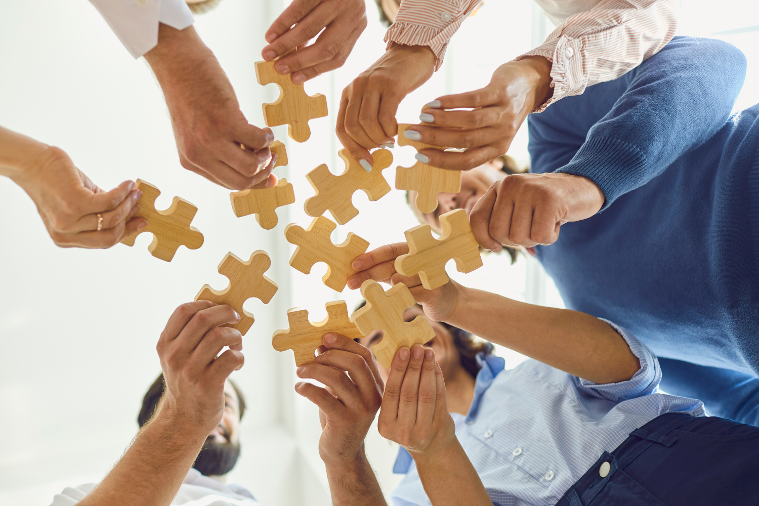 Multiple people are working together, holding and connecting wooden puzzle pieces in an upward motion, symbolizing teamwork and collaboration.