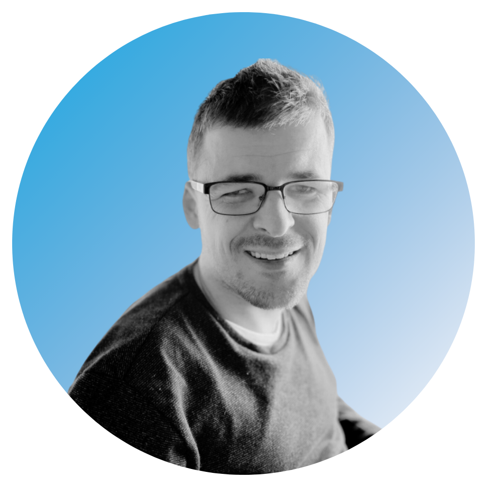 Black and white headshot of a smiling man with glasses, styled hair, wearing a dark shirt, against a blue background.