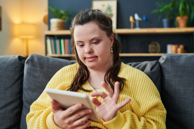 A woman in a yellow sweater sitting on a gray couch, looking at her smartphone in a cozy living room with books, plants, and decor in the background.