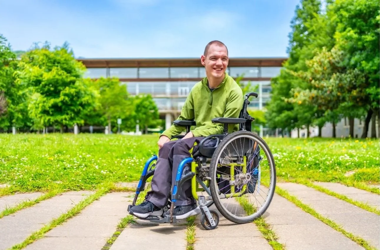 A smiling man in a wheelchair outdoors on a sunny day, with green trees and a modern building in the background.