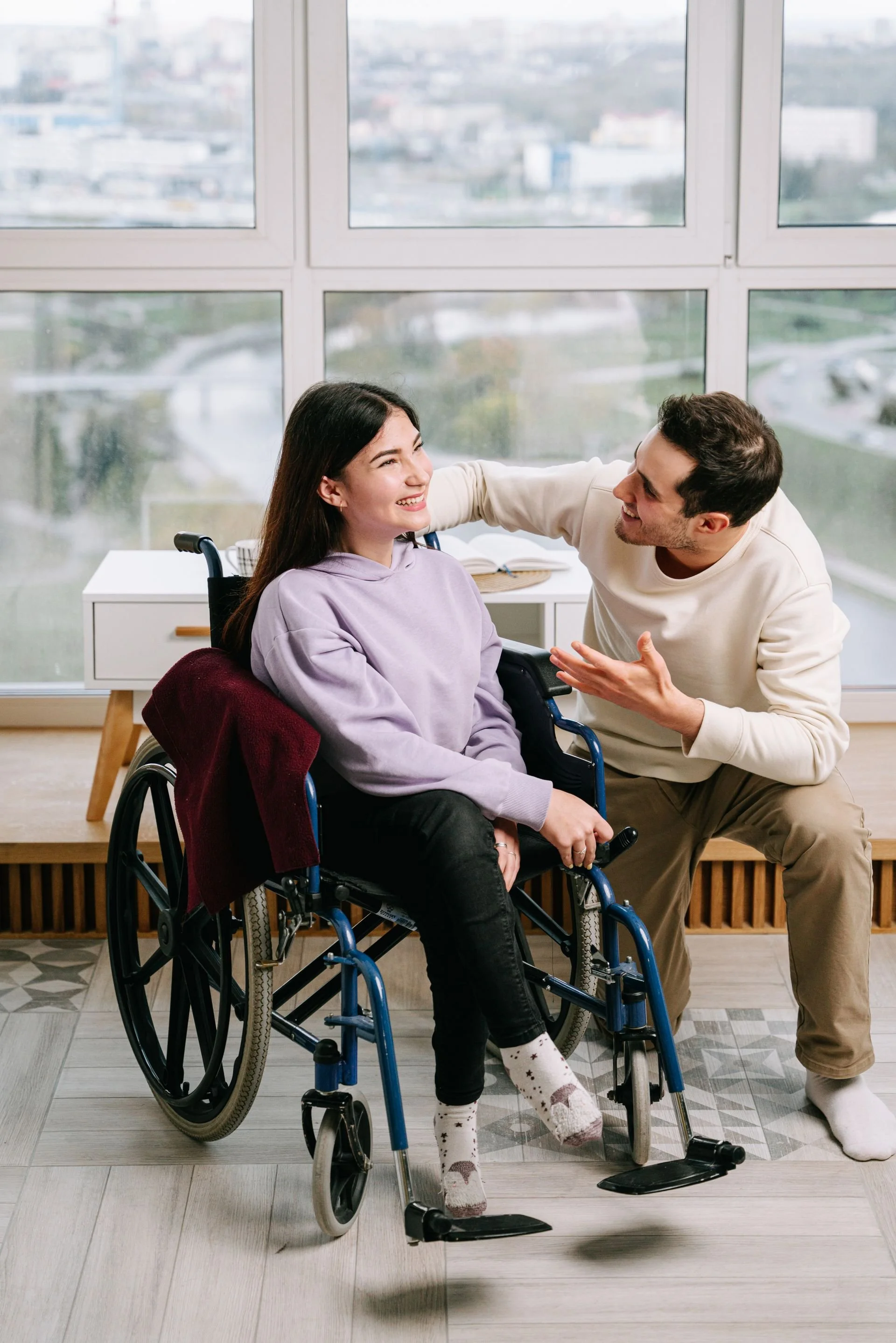 A young woman in a wheelchair smiling and looking at a man who is kneeling beside her, engaging in conversation. They are indoors with large windows and a city view in the background.