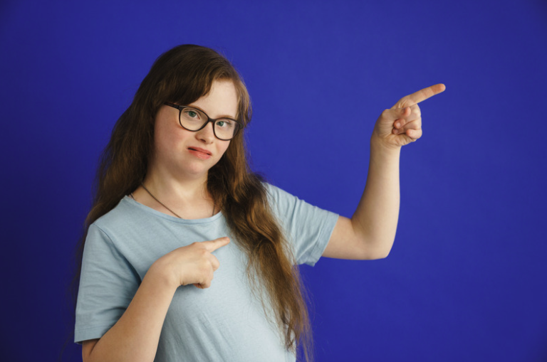 A young woman with long brown hair and glasses pointing to her right with her left hand and to her chest with her right hand against a plain blue background.