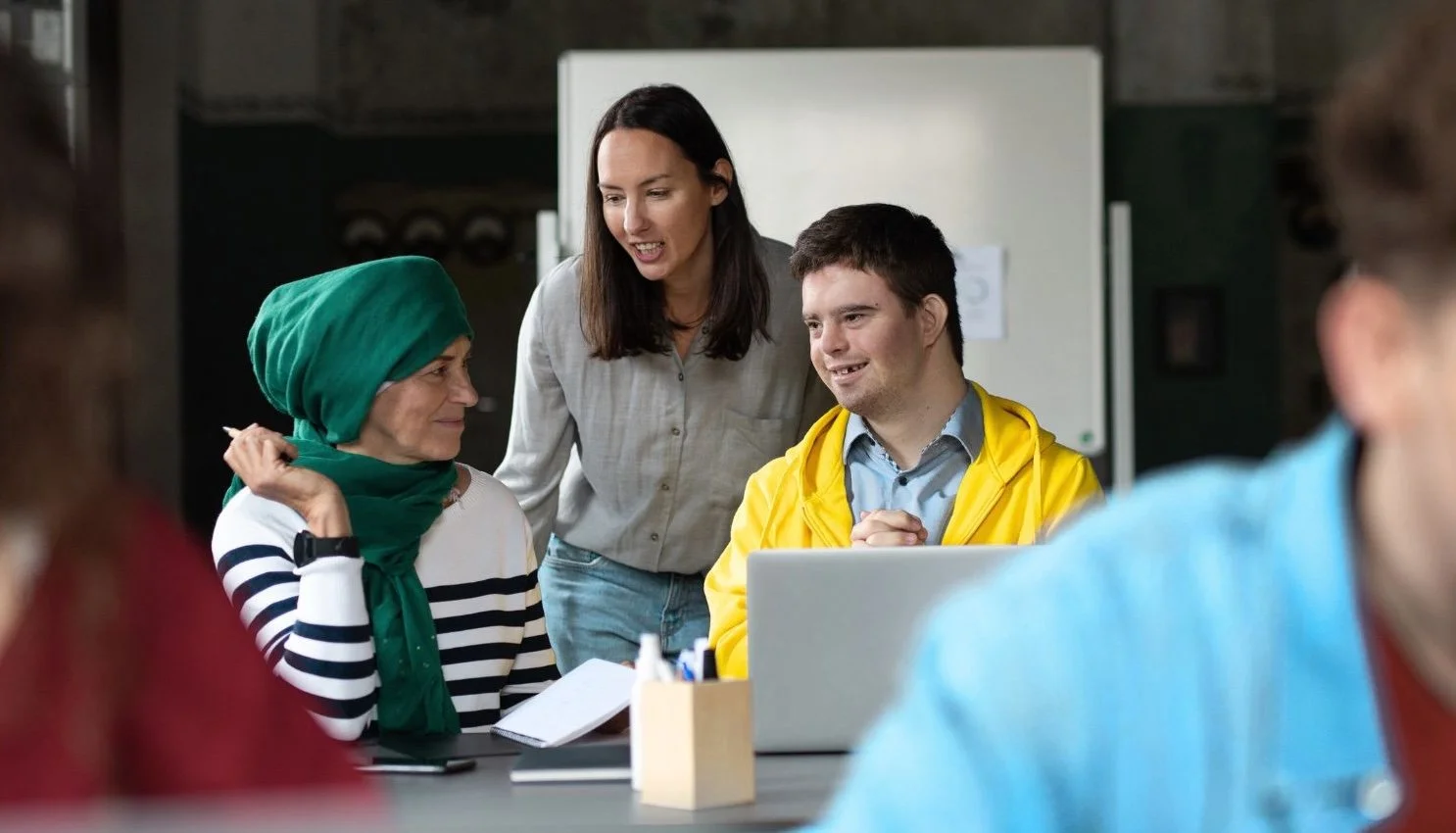 A group of diverse people having a meeting in a casual office setting, with a woman wearing a green headscarf and striped shirt talking to a young man in a yellow jacket, while a woman with dark hair observes.