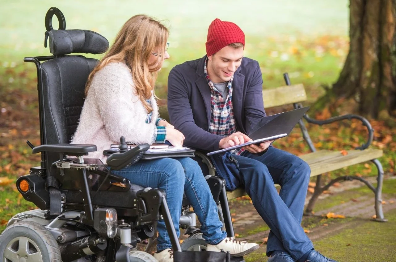 A young man with a red beanie and a young woman in a pink sweater are sitting on a park bench outdoors, looking at a laptop and a notebook together. An electric wheelchair is beside them.