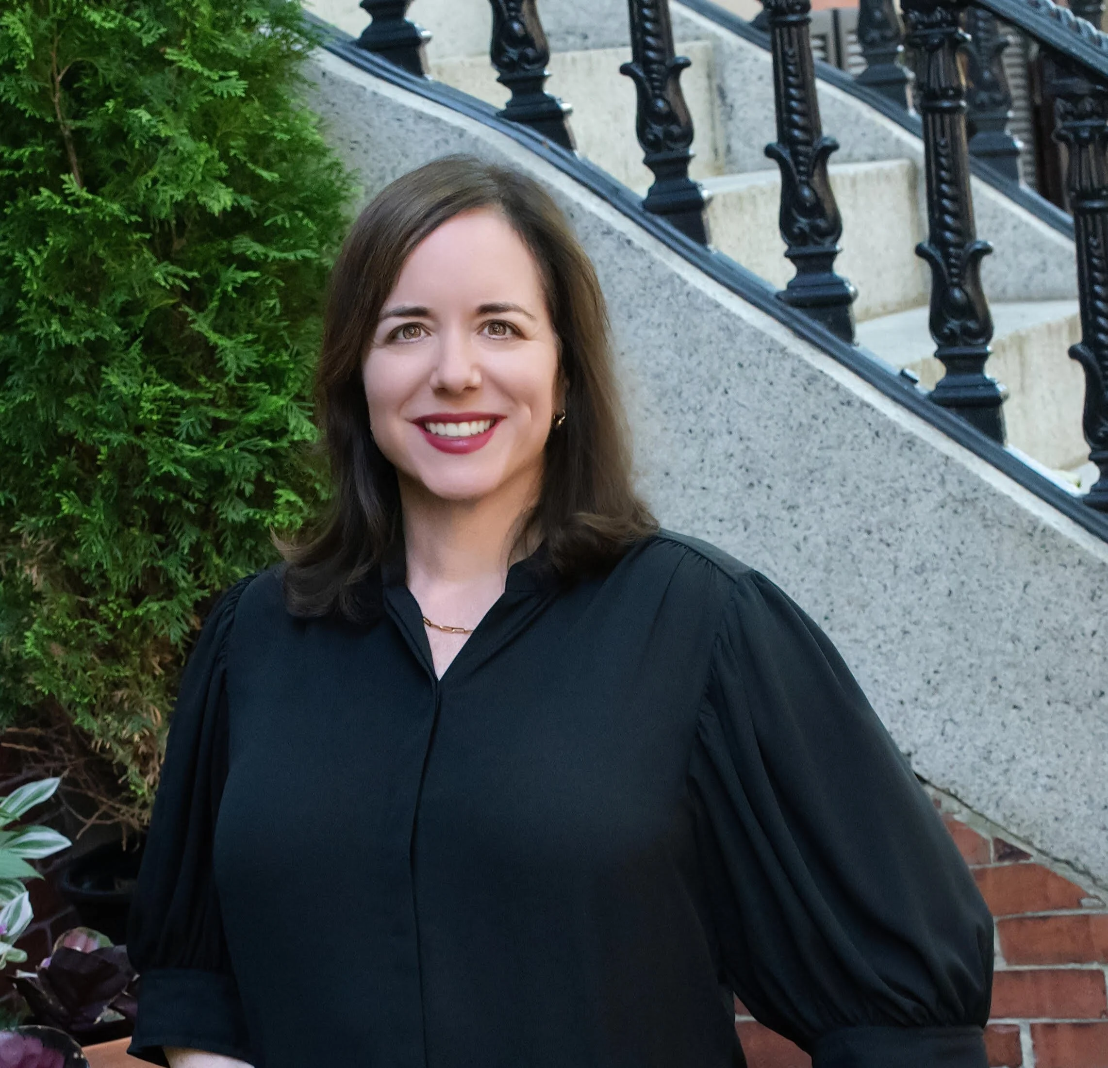 A woman with dark brown hair and light skin smiling outdoors, wearing a black top and gold necklace, in front of a green bush and a stairway with black wrought-iron railings and stone steps.