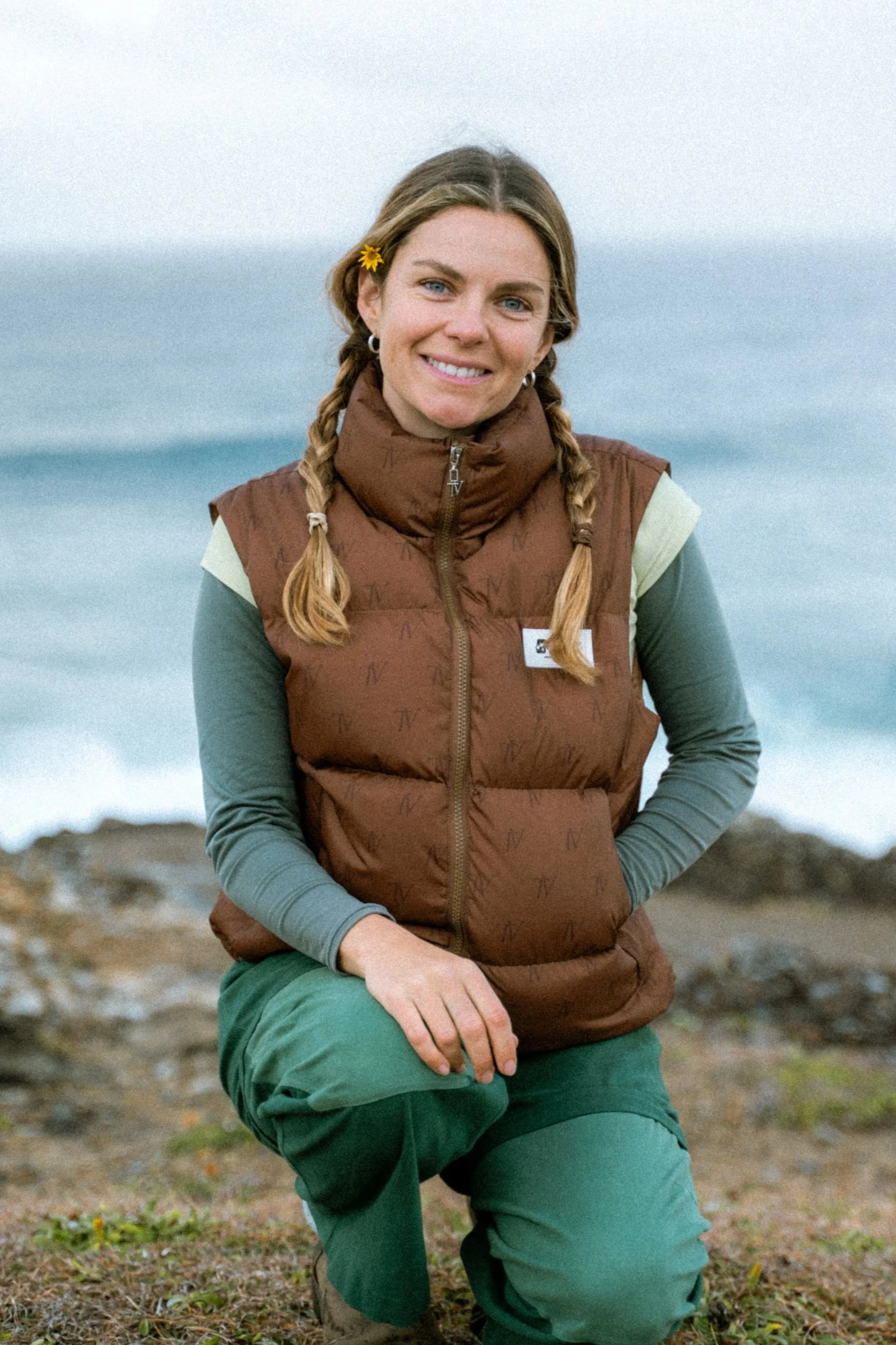 Woman kneeling on rocky beach, smiling, wearing brown vest, green pants, and a yellow flower in her hair, with ocean in background.