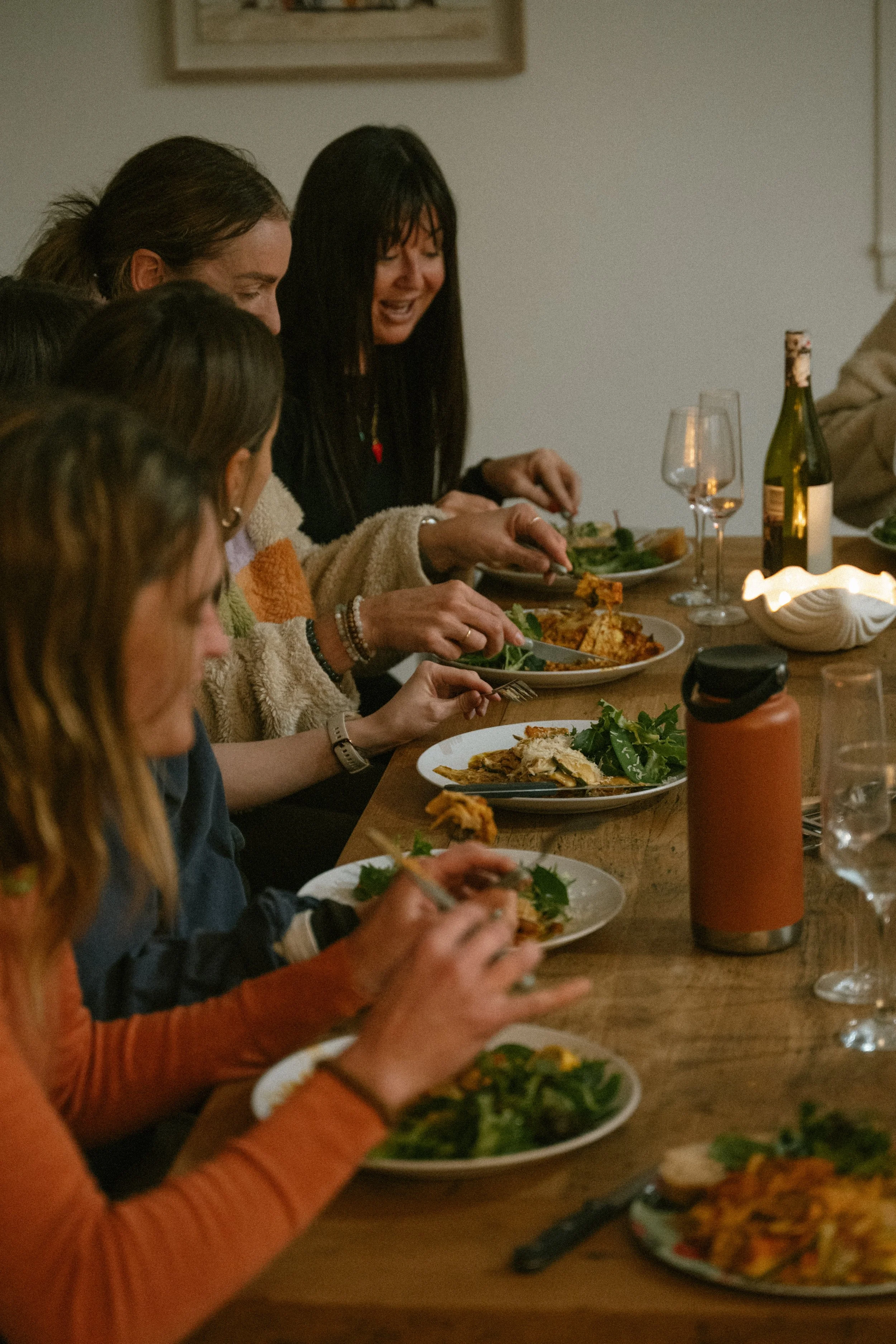 A group of people sitting around a dinner table, enjoying a meal with plates of food, wine bottles, glasses, and a lit candle.