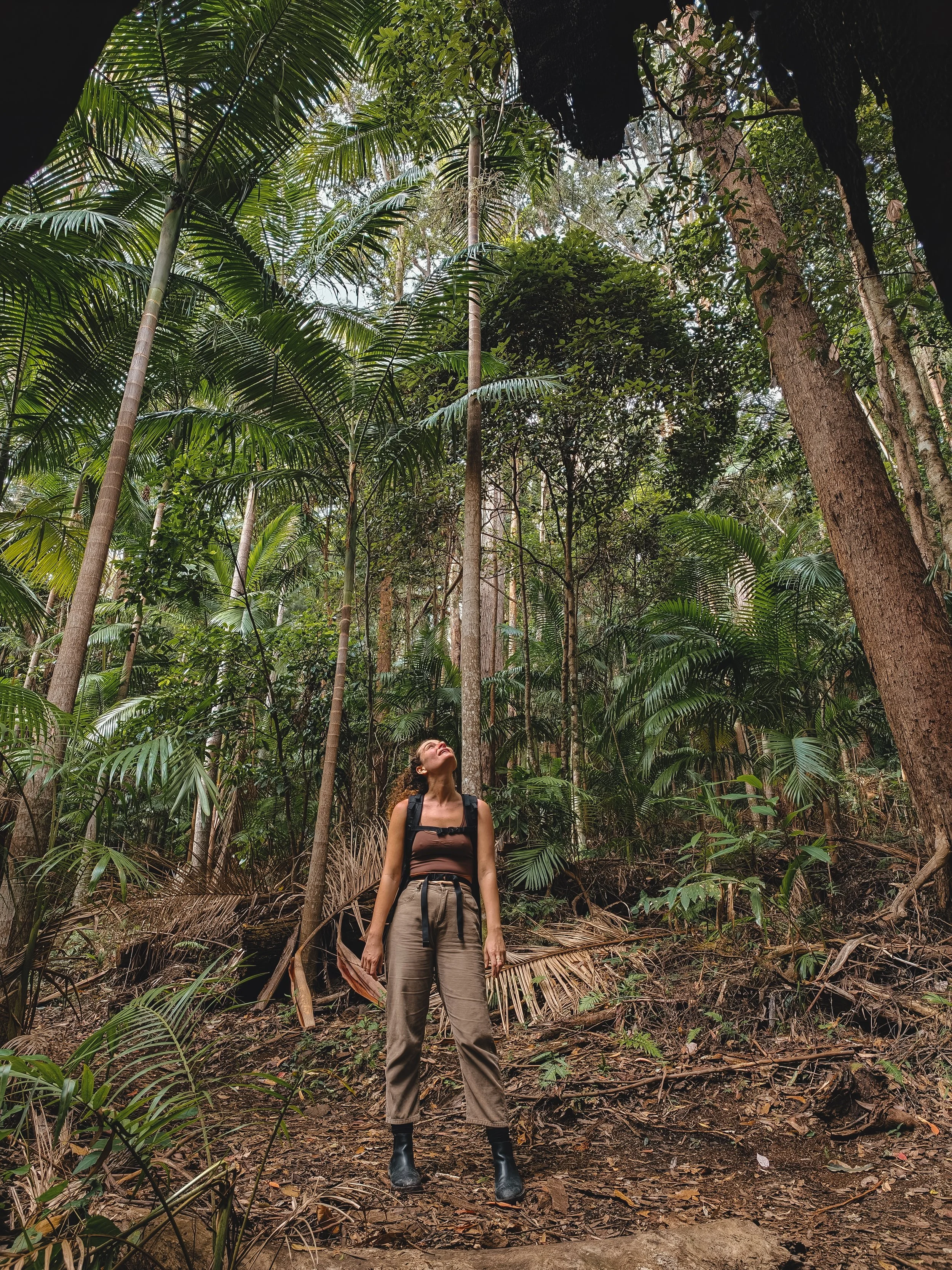 A woman with glasses, wearing a brown tank top, beige pants, and black boots, stands in a lush green jungle looking upwards.