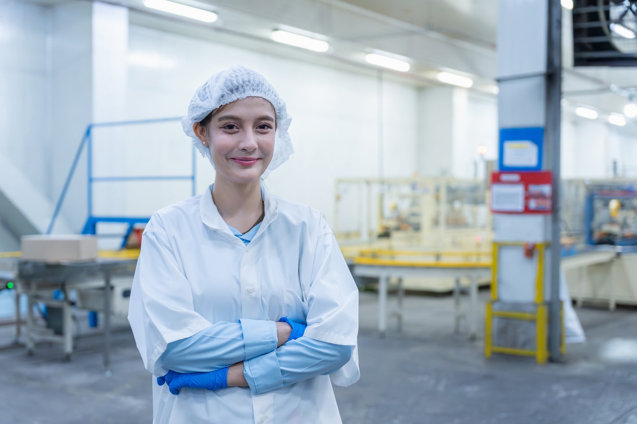 A female worker wearing a hairnet, white lab coat, and blue gloves standing in a factory or manufacturing facility with equipment and assembly lines in the background.