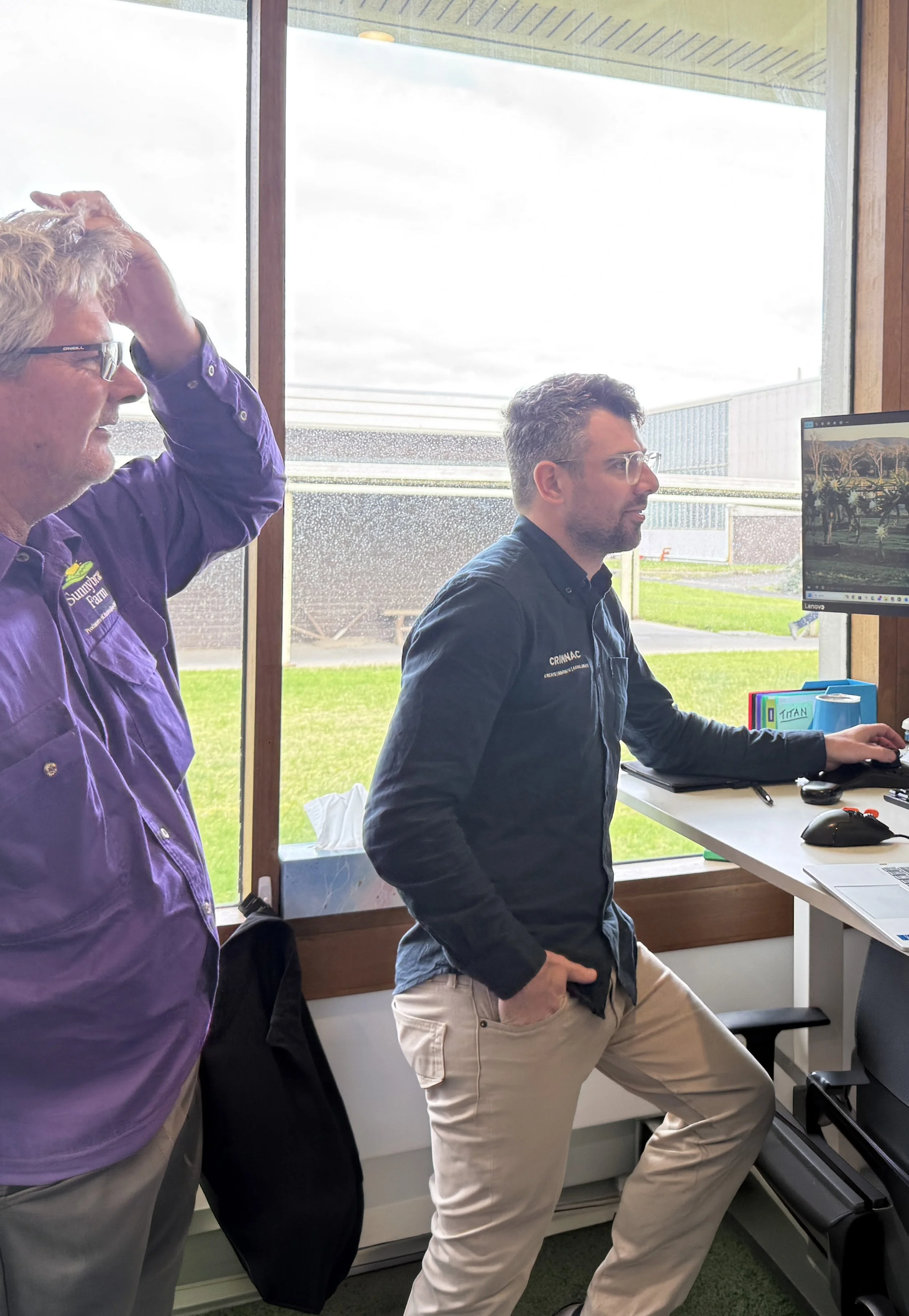 Two men are standing in front of a computer screen inside a room with large windows showing an outdoor industrial area. One man is wearing a purple shirt and the other is in a dark shirt and beige pants. The man in purple is touching his head, the other man is sitting with his right hand on his hip, looking at the computer monitor.