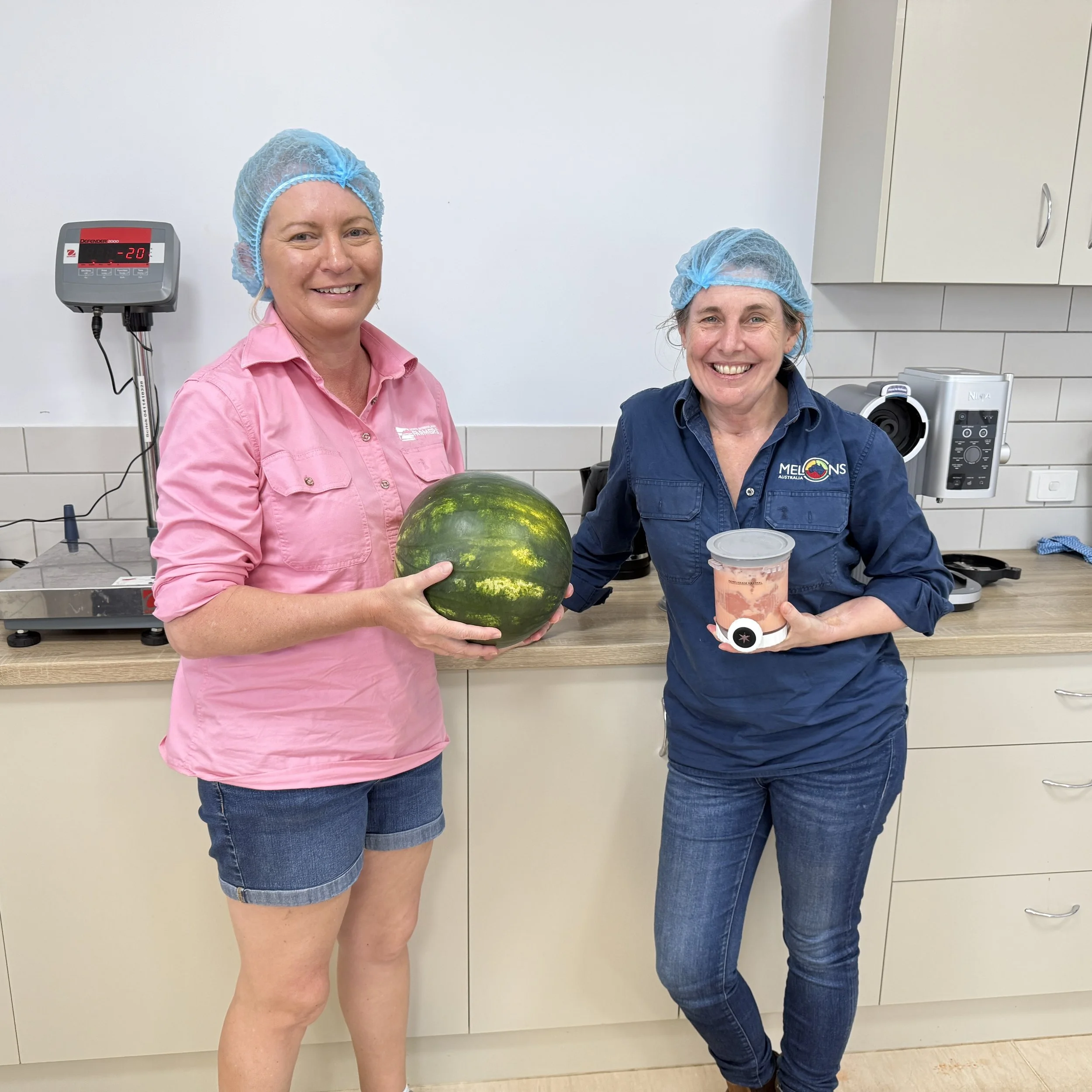 Two women wearing blue hair nets smiling in a medical or laboratory setting. One woman in a pink shirt holds a watermelon, and the other woman in a dark blue shirt holds a container. They are standing in front of a white wall with medical equipment.