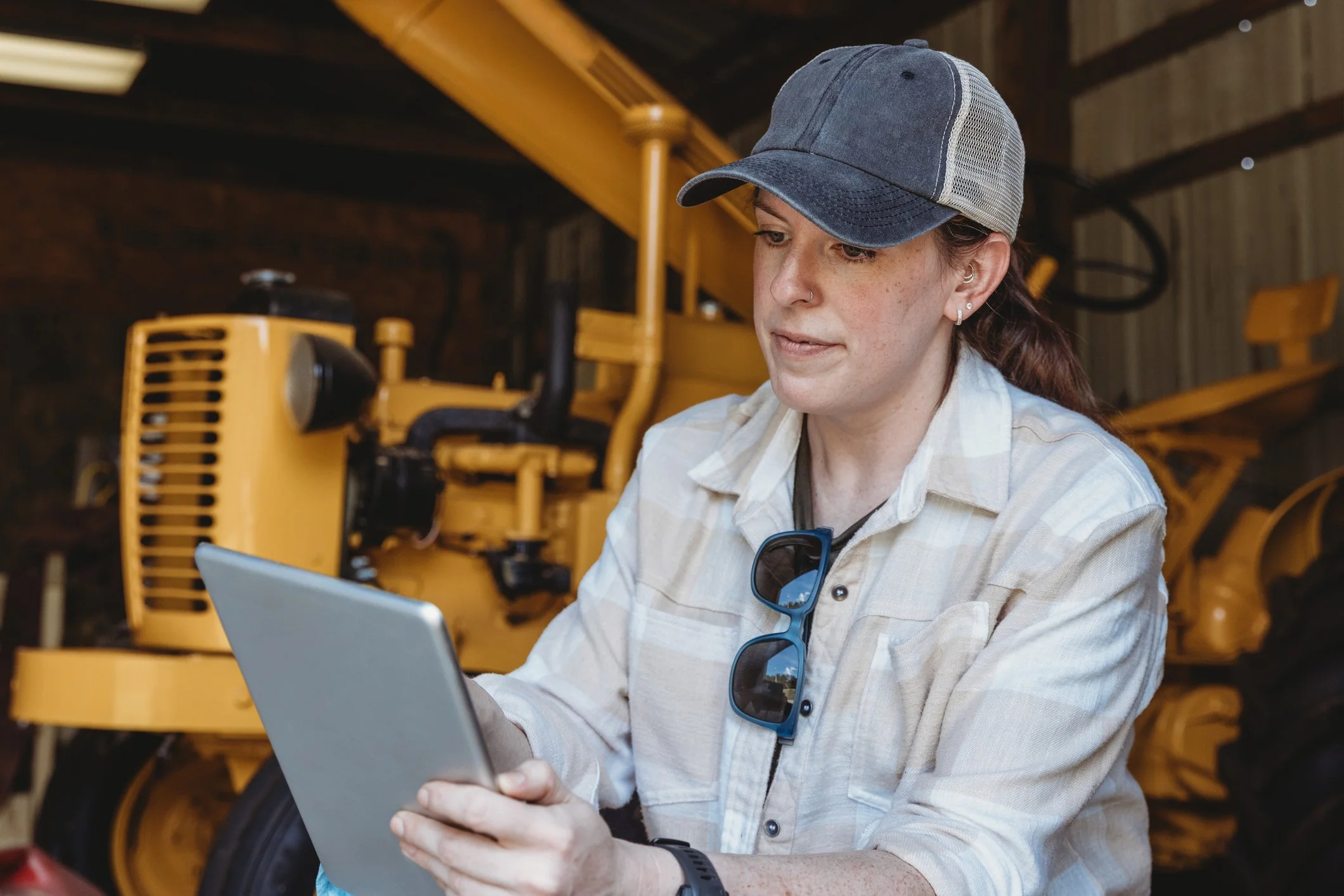 A woman wearing a denim baseball cap, earrings, and a plaid shirt is sitting indoors, looking at her laptop and holding a smartphone. A yellow piece of farm machinery is visible in the background.