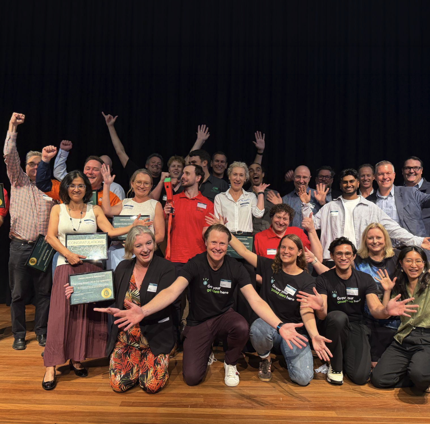 A large group of people on a stage celebrating, smiling, and waving at the camera, with some holding certificates and awards.