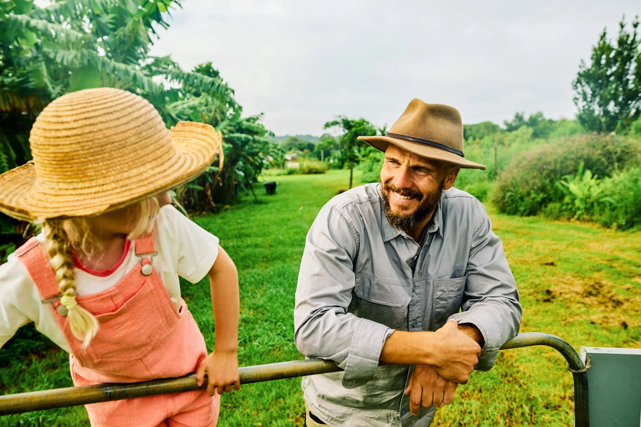 A man and a young girl with braided hair, both wearing hats, are smiling and talking outdoors in a lush green garden or park setting.