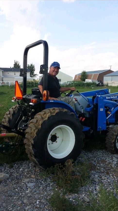 A man sitting on a blue New Holland tractor with large, muddy tires on a rural property with houses and barns in the background.