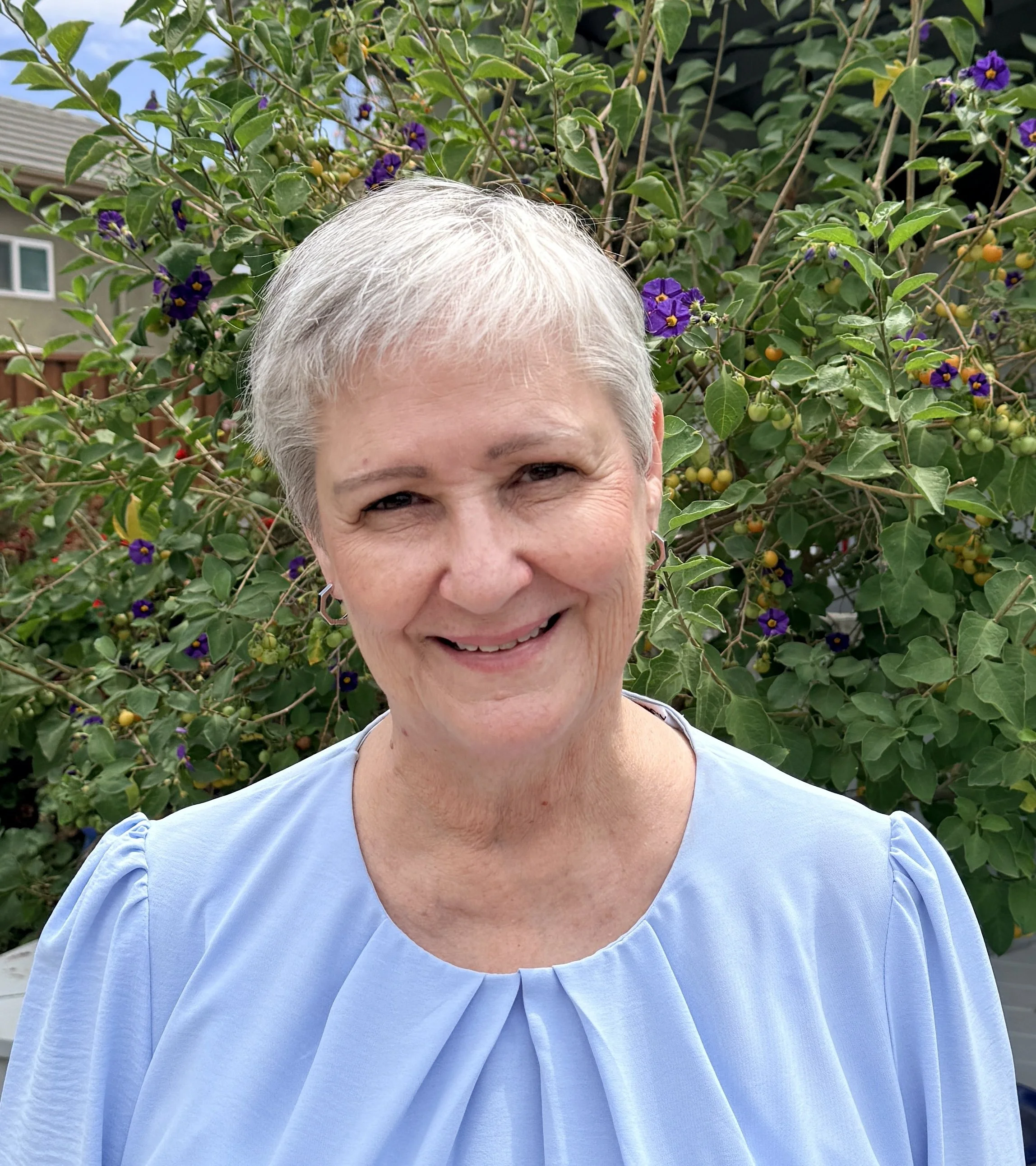 A woman with short gray hair smiling in front of a lush green bush with purple and yellow flowers.