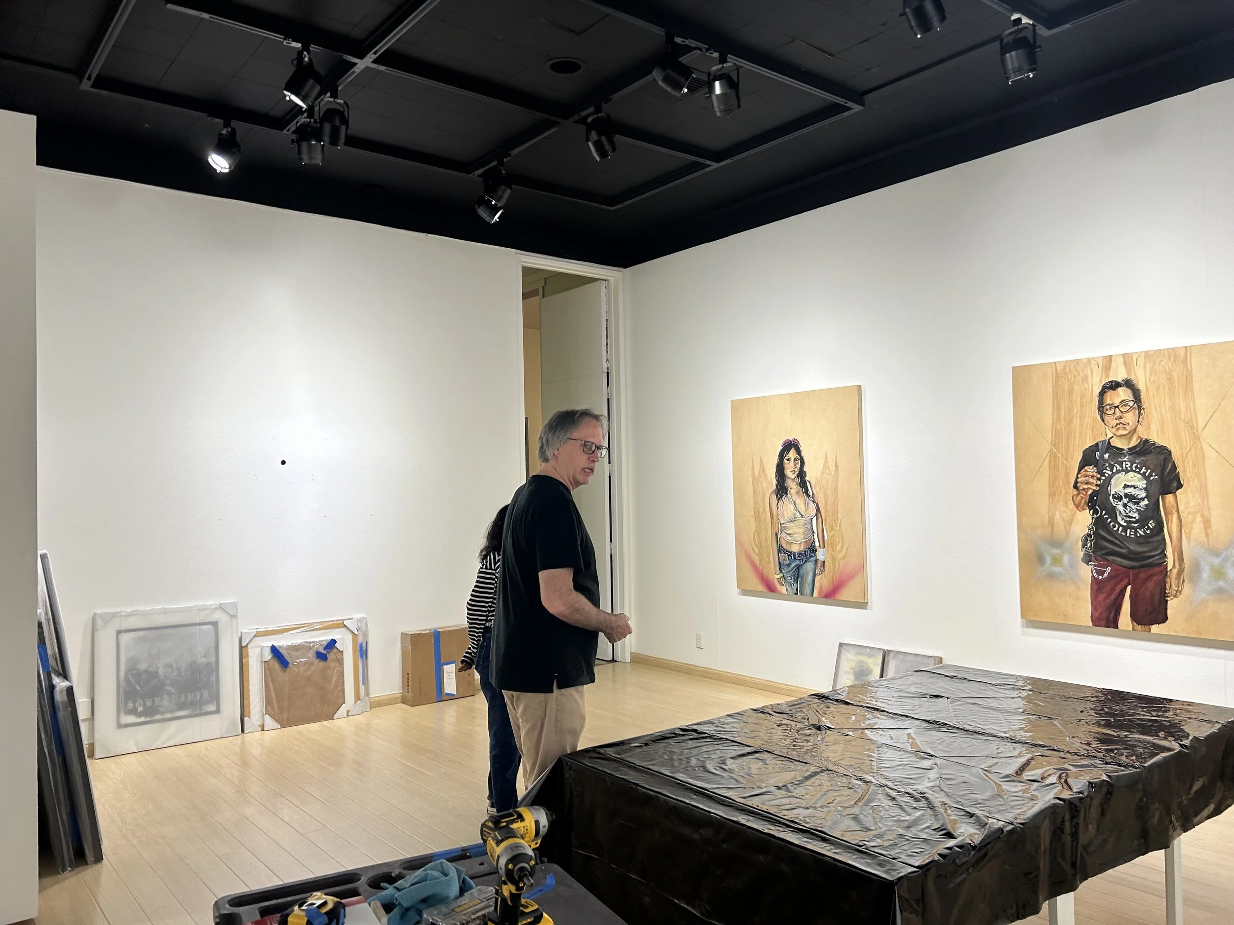 Two people in a gallery or studio surrounded by artwork and installation supplies, with paintings on the wall and tools on a table covered in black material.
