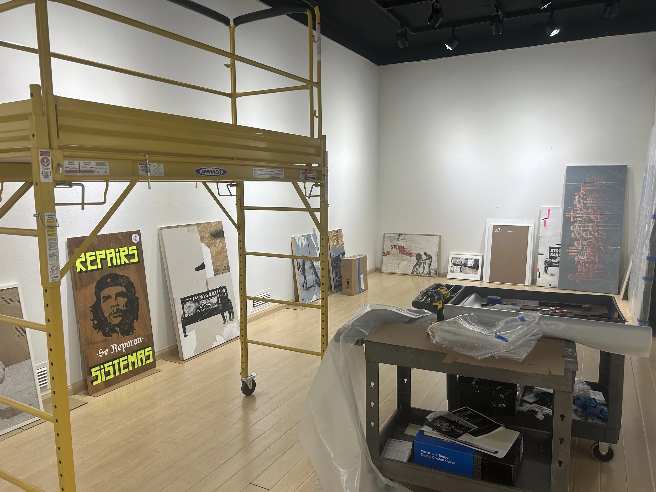 Art supplies and framed posters arranged in an art studio or gallery space. A yellow scaffolding is present, and the room has white walls and a light wood floor. Some tools and packaging are on a nearby table.