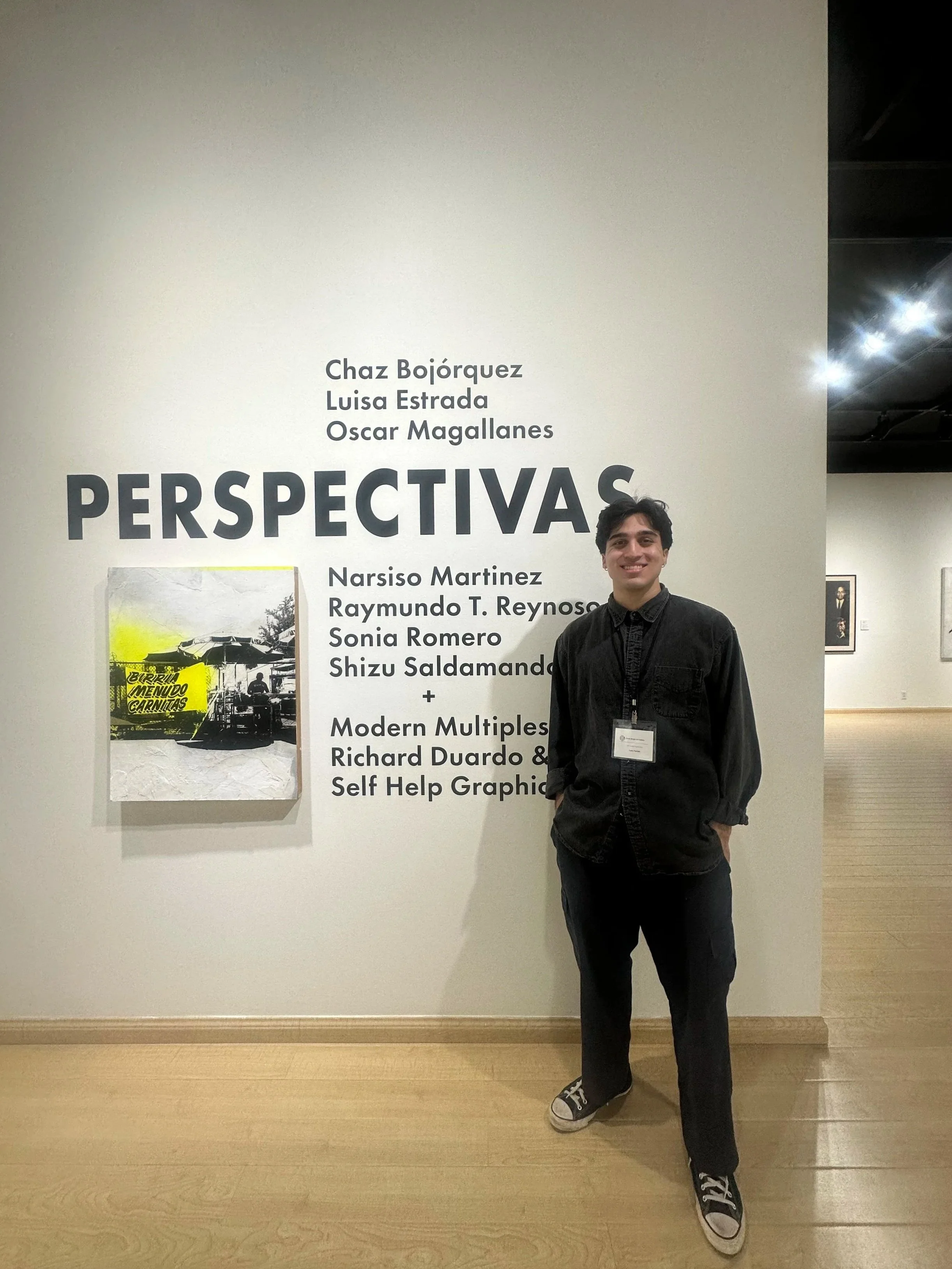 Young man smiling and standing with hands in pockets in front of a white exhibition wall with text and artwork displayed.
