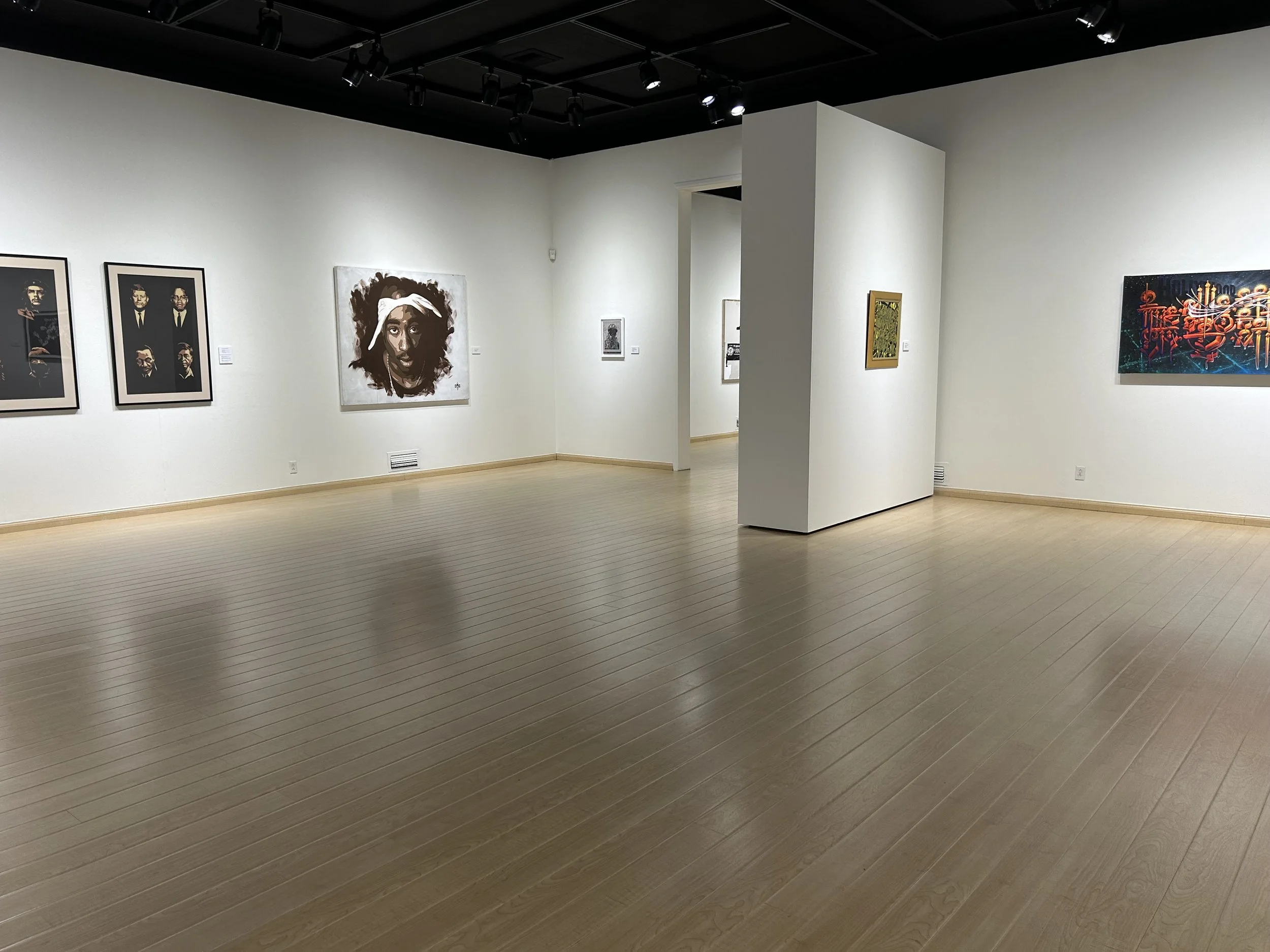 Art gallery with paintings on white walls and wooden flooring, featuring portraits and abstract artwork.