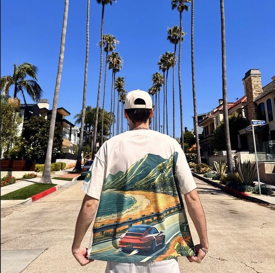 Person wearing a white t-shirt with a landscape and car design on the back, walking down a tree-lined street with palm trees and houses, under a clear blue sky.