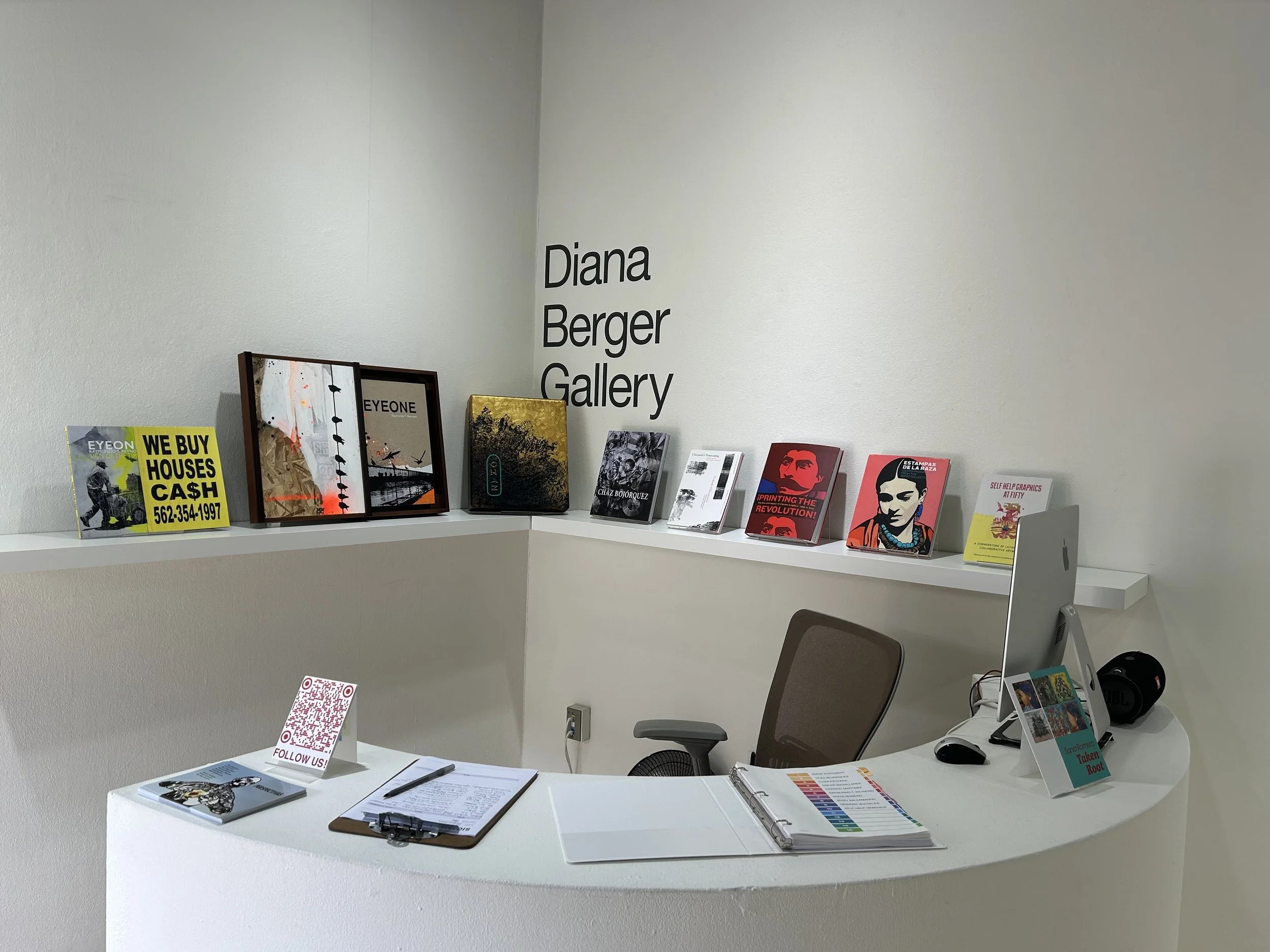 An art gallery reception area with a white wall displaying black text reading 'Diana Berger Gallery' and various art books and printed materials arranged on a white counter and shelves, including a yellow sign with black text and yellow sign with black text, and a brown office chair and a small table with a colorful color chart, QR code, and a few papers.