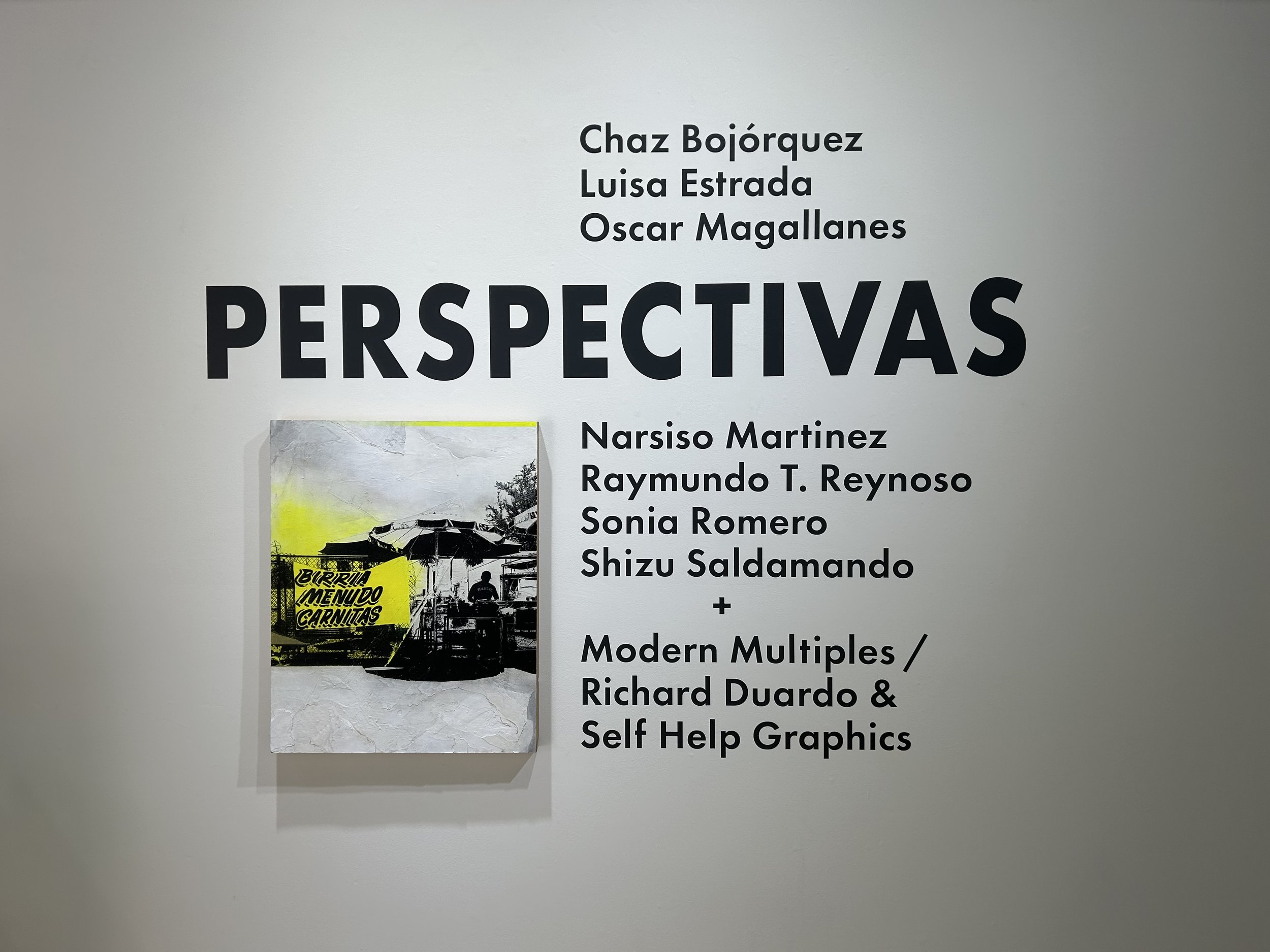 White wall with black text displaying names of artists and a list of art projects, with a poster partially visible at the bottom left corner.