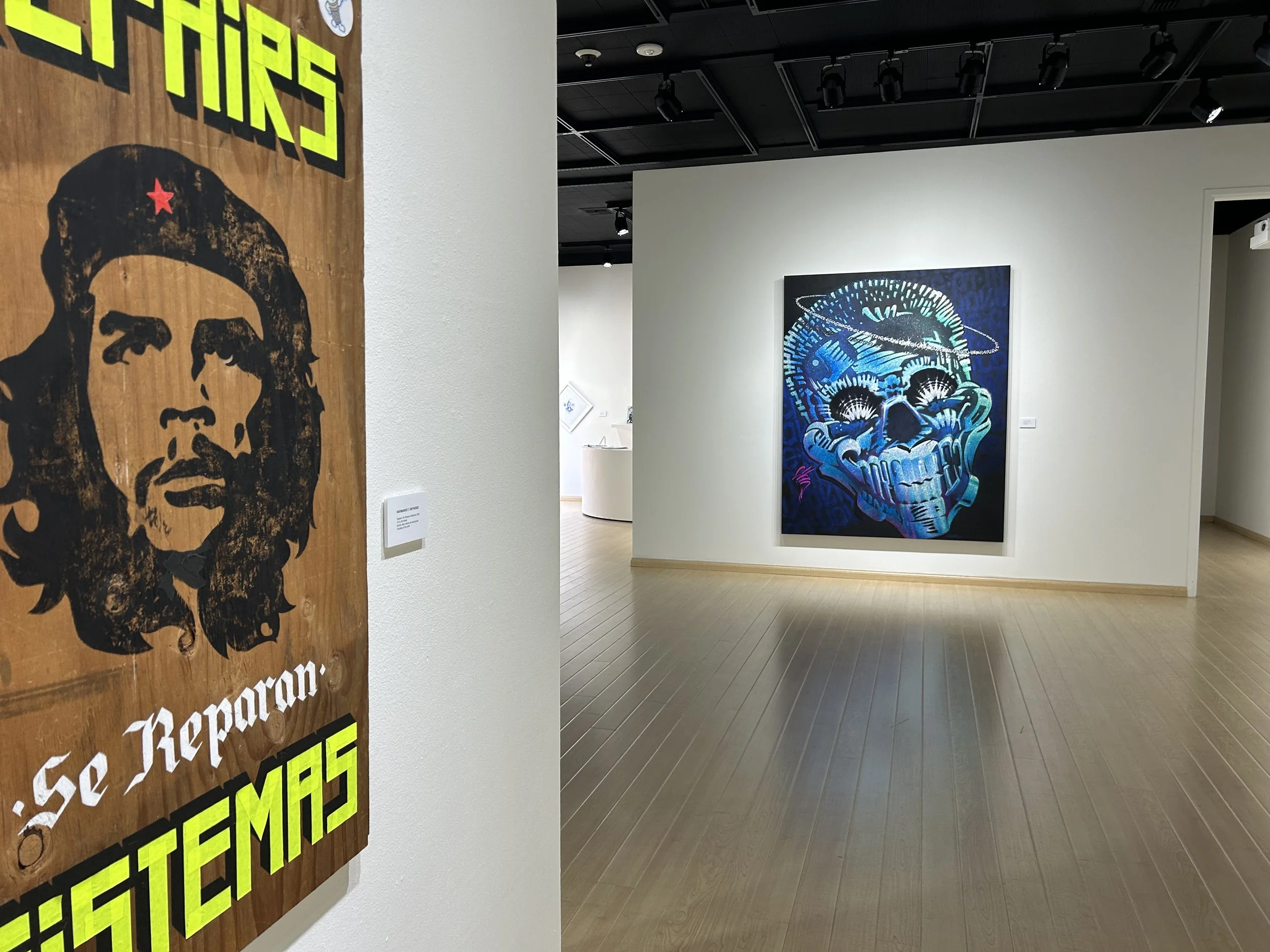 View of an art gallery with a large colorful painting of a human skull and a smaller painting of a person in the background, with walls and ceiling lights.