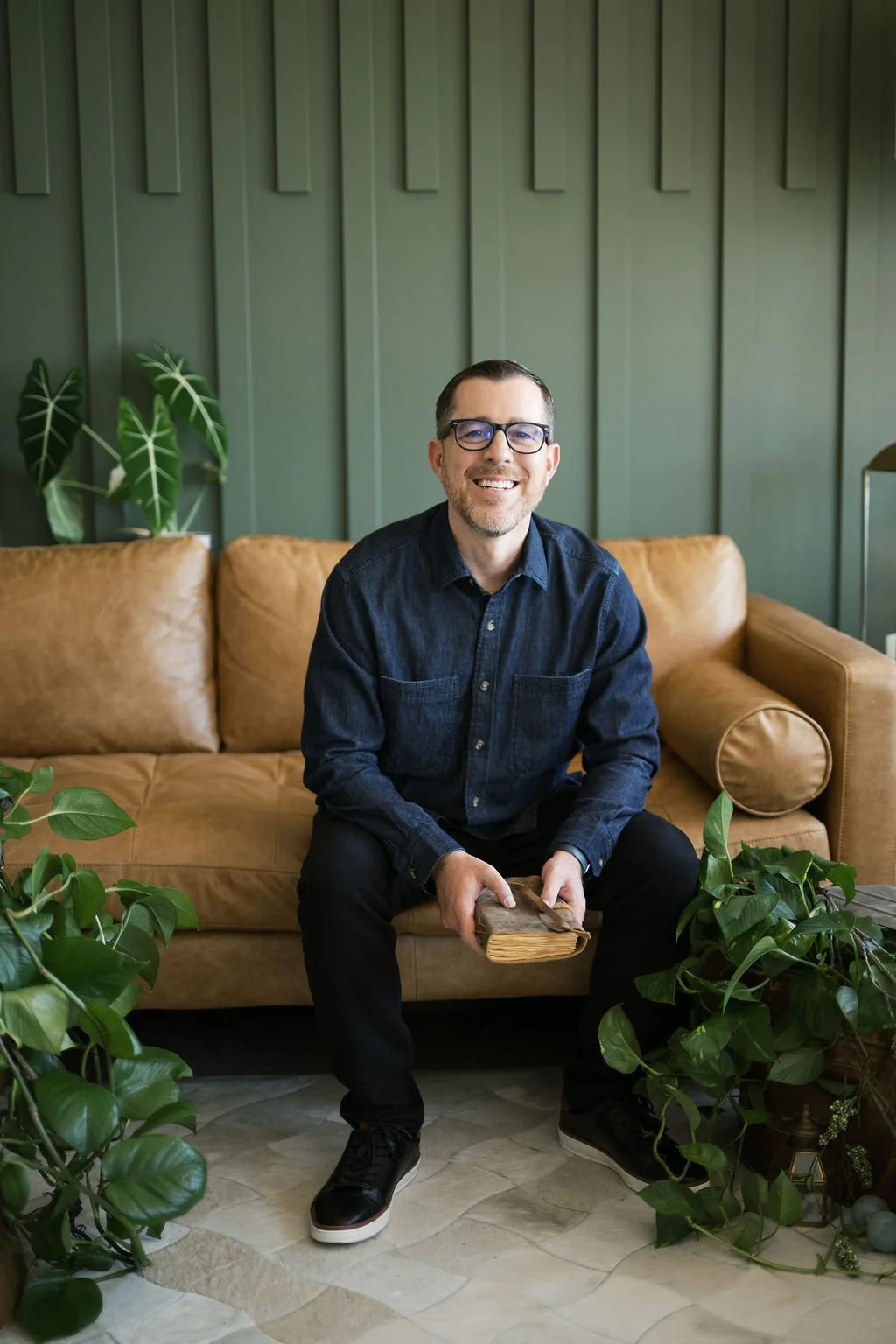 A smiling man with glasses, wearing a dark denim shirt and black pants, sitting on a tan leather sofa, holding a book, with indoor green plants around him and a green paneled wall behind.