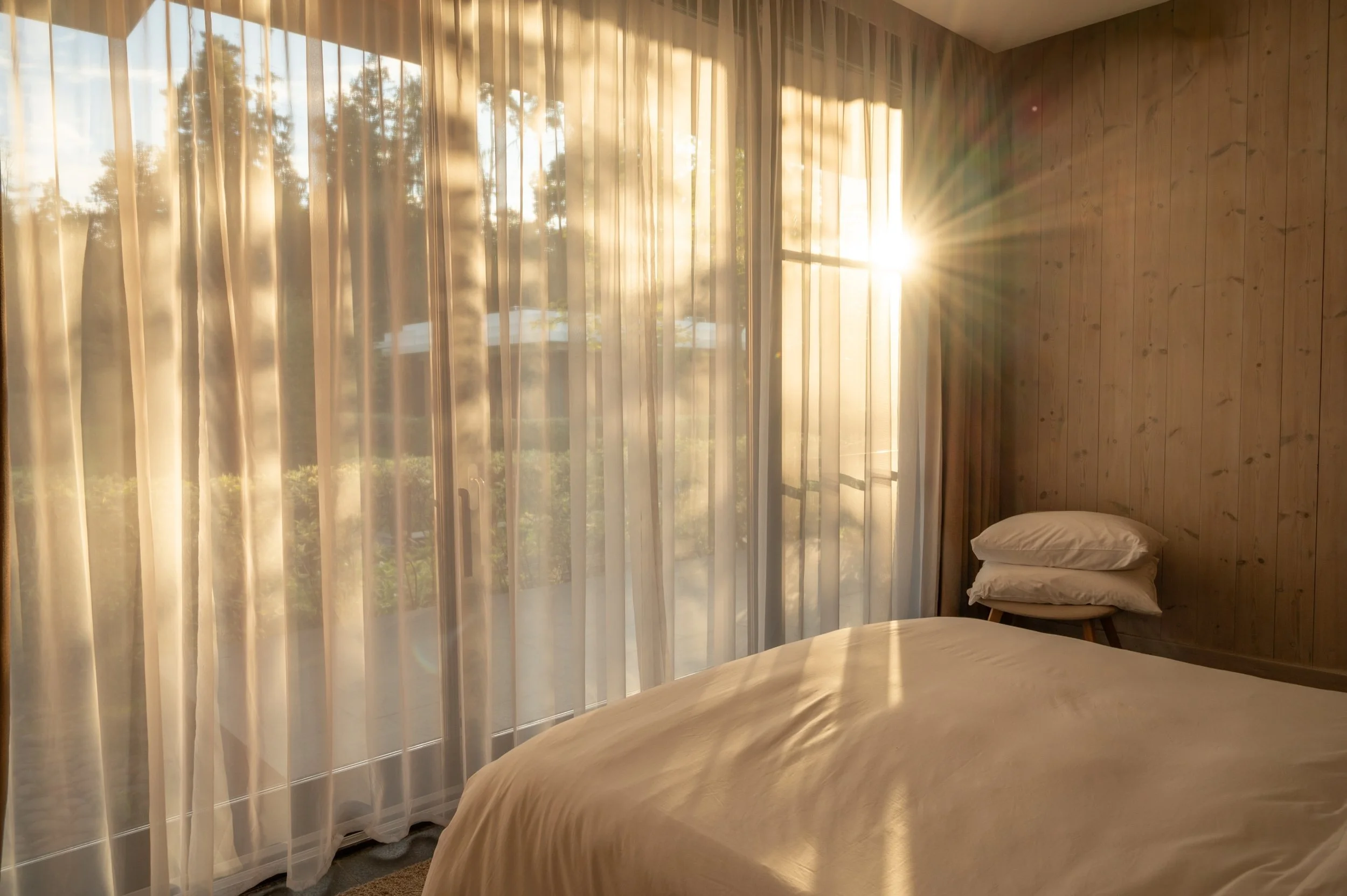 Sunlight streaming through sheer curtains in a bedroom, illuminating a made bed with pillows and wooden-paneled walls.