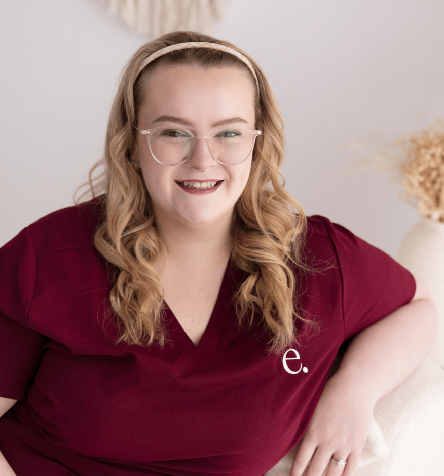 A woman with long, wavy blonde hair wearing glasses and a maroon shirt with a white 'e.' logo, sitting in a neutral indoor setting, smiling at the camera.