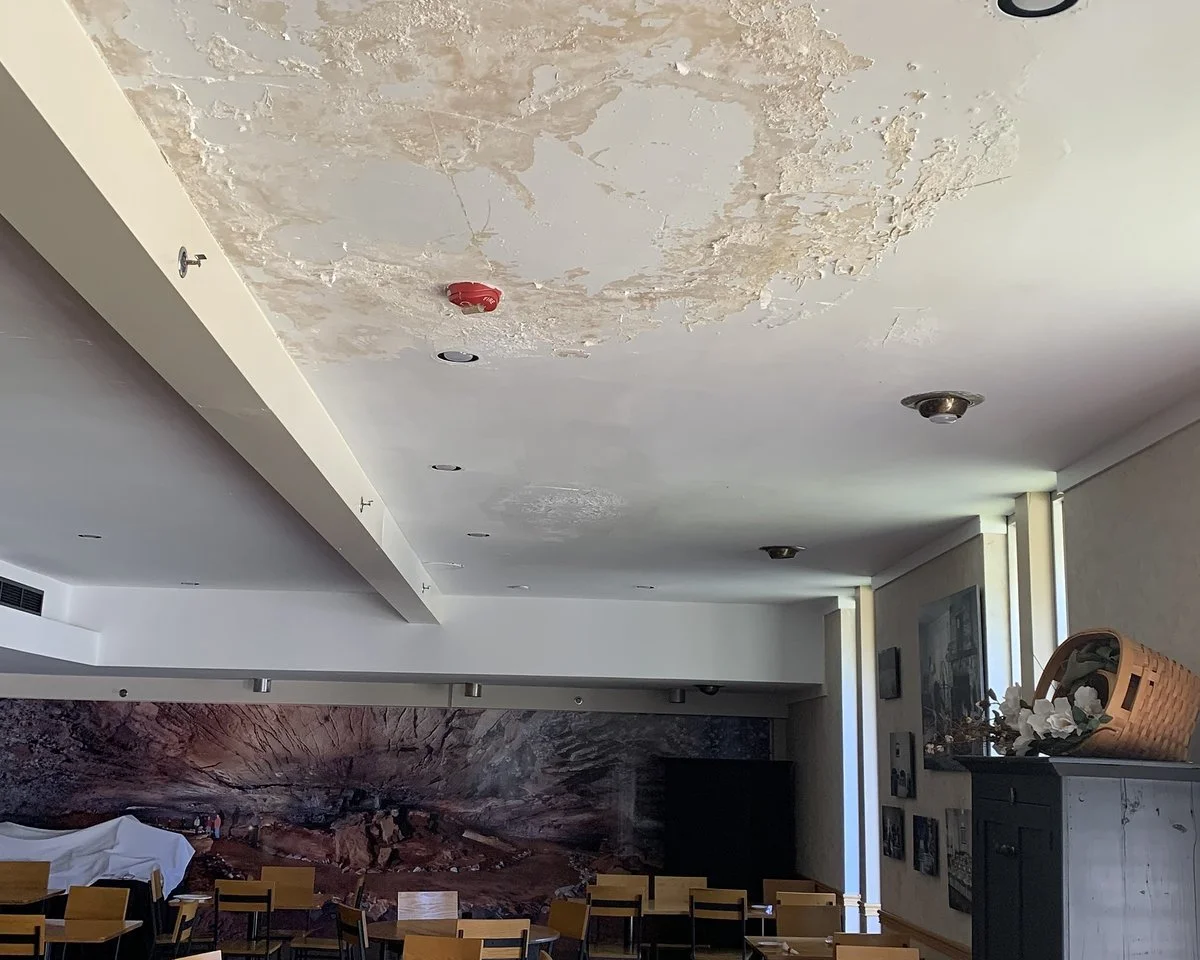 Peeling, water-stained ceiling from a roof leak above a commercial dining room with rows of empty tables and chairs.