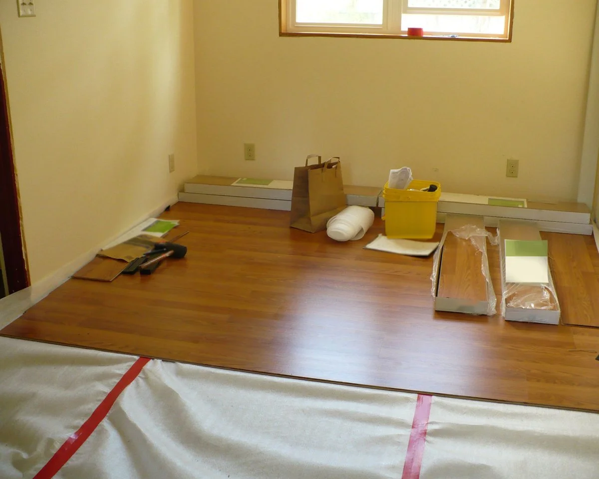 A half-installed wood-look laminate floor in an empty room, with underlayment still showing, flooring boxes, a hammer and installation supplies on the boards.