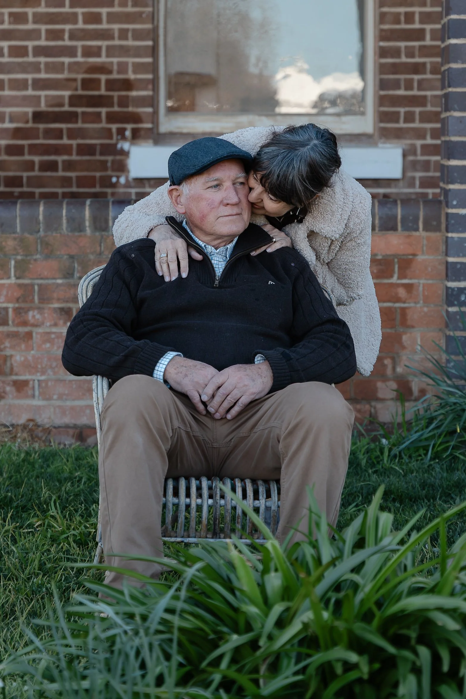 An elderly man sitting on a chair outdoors in front of a brick house, being closely embraced and comforted by a woman leaning in to him, with greenery and plants in the foreground.