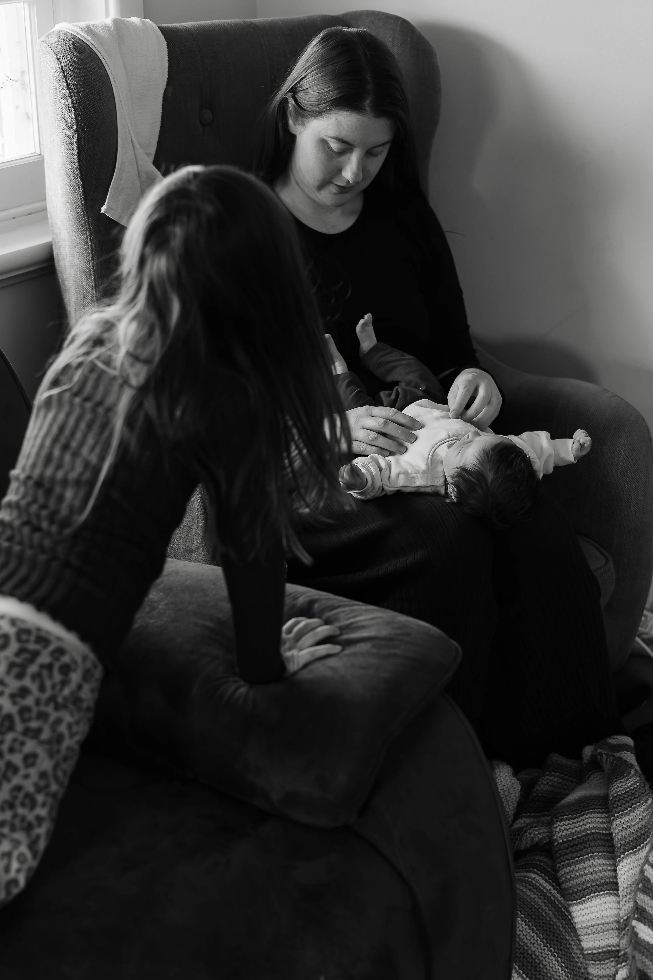 A woman with long dark hair and freckles sitting on a couch, looking down at a baby lying on her lap. The baby appears to be resting or sleeping, with dark hair, wearing a light-colored outfit. An older child with long, hairy arms and leopard print s