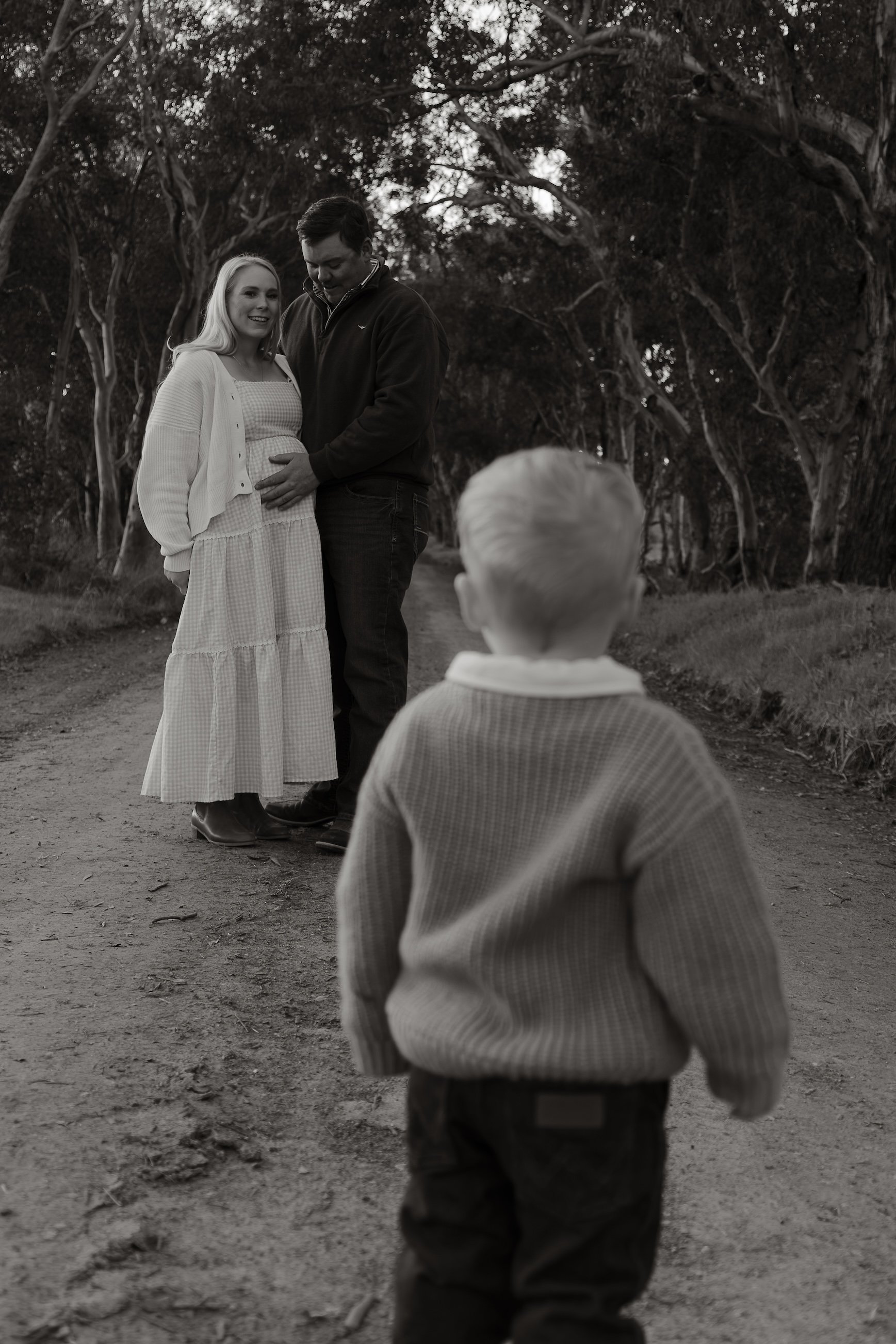 A pregnant woman and a man standing together in a wooded outdoor area, with a young boy observing them from behind in the foreground.