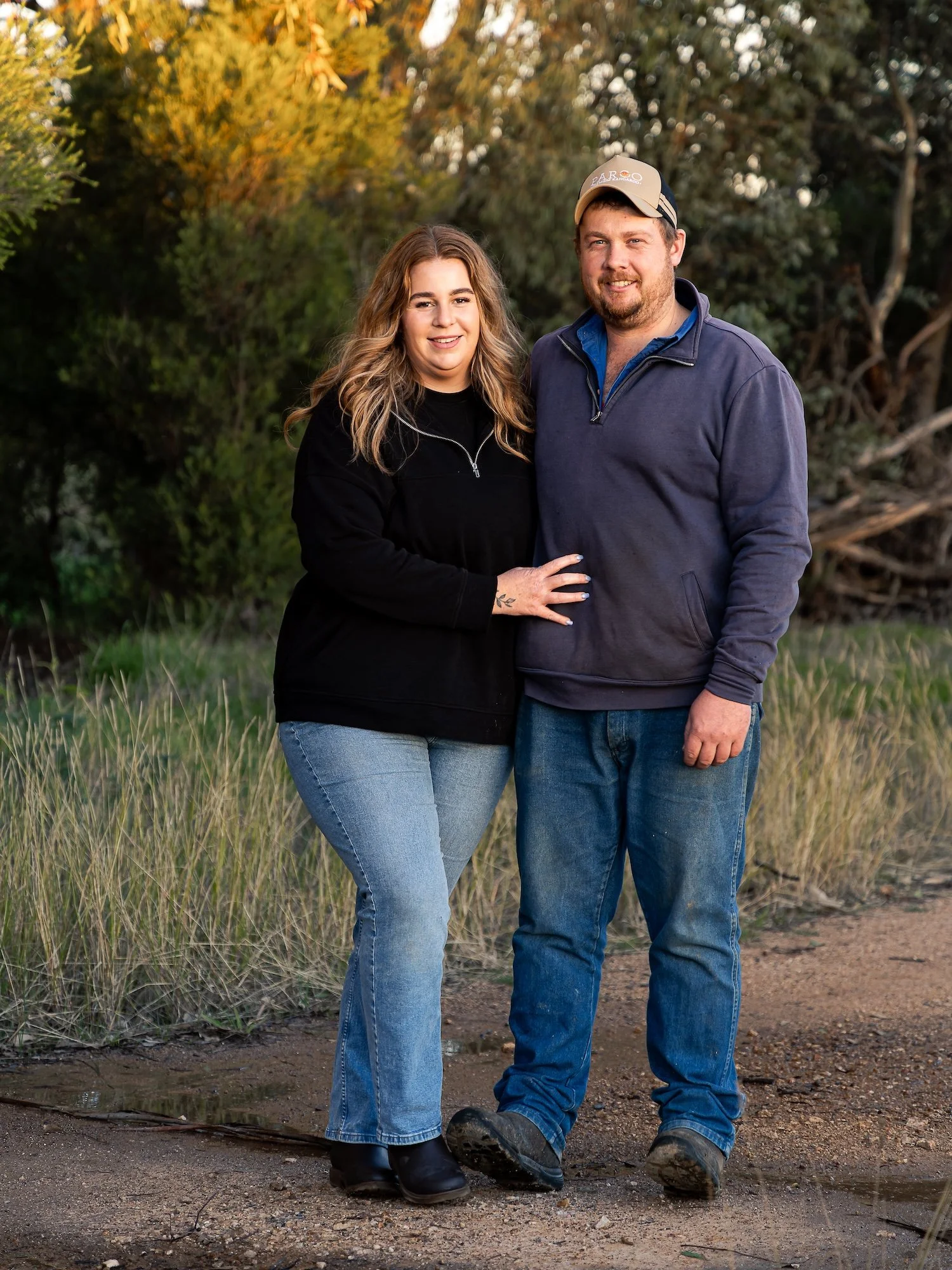 A man and woman standing together outdoors on a dirt path with trees in the background during autumn.
