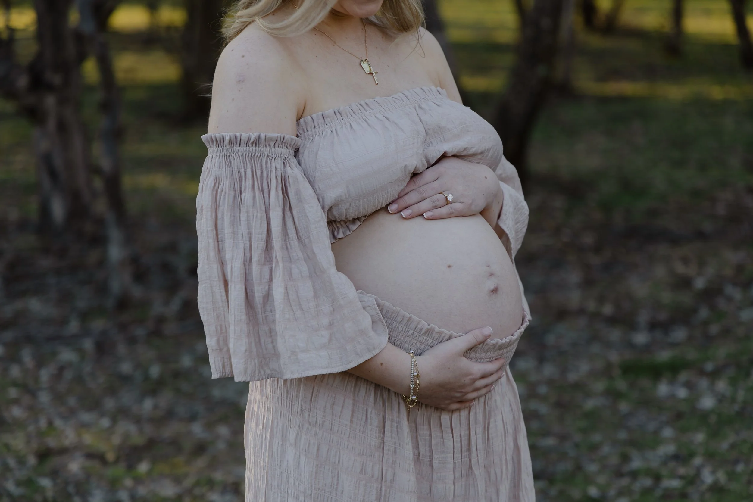 A woman with blonde hair wearing an off-the-shoulder beige dress holding her pregnant belly outdoors in a wooded area.