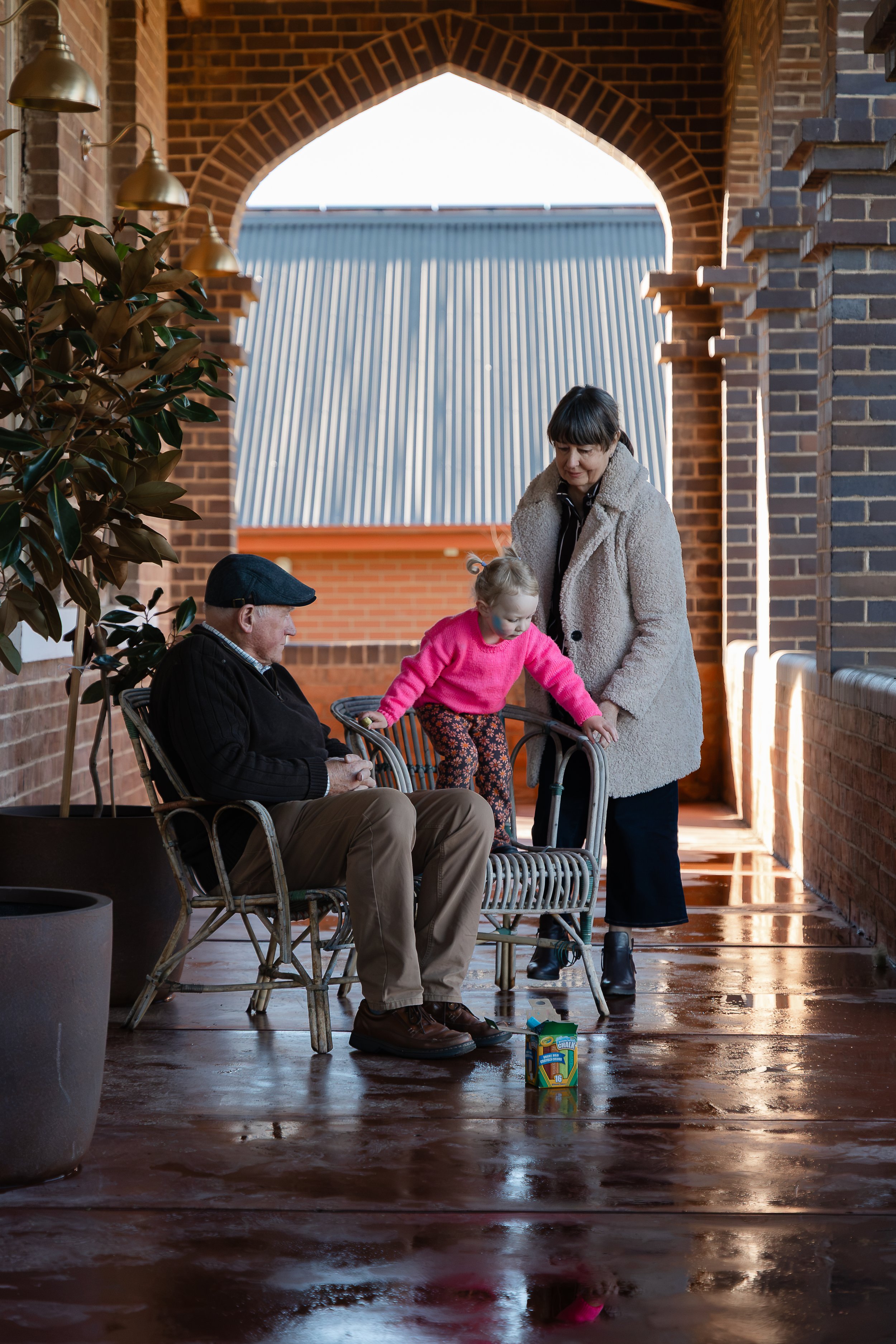 A woman, a young girl, and an older man are sitting on wicker chairs on a covered porch with brick walls. The woman and girl are standing near each other, and the girl is reaching for a juice box on the wet floor. The man is seated with his head slig