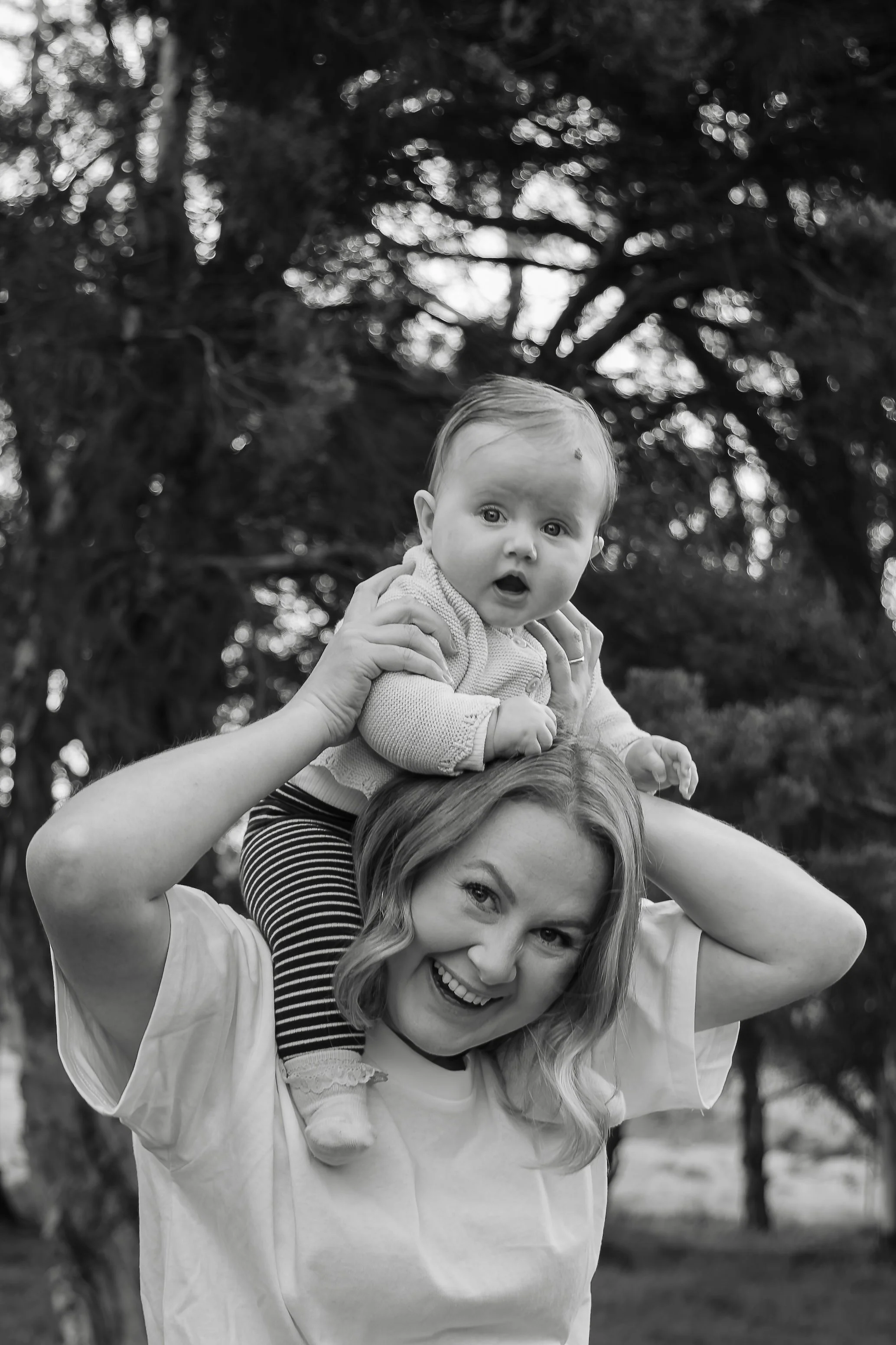 A smiling woman with wavy hair carrying a baby on her shoulders outdoors, with trees in the background.