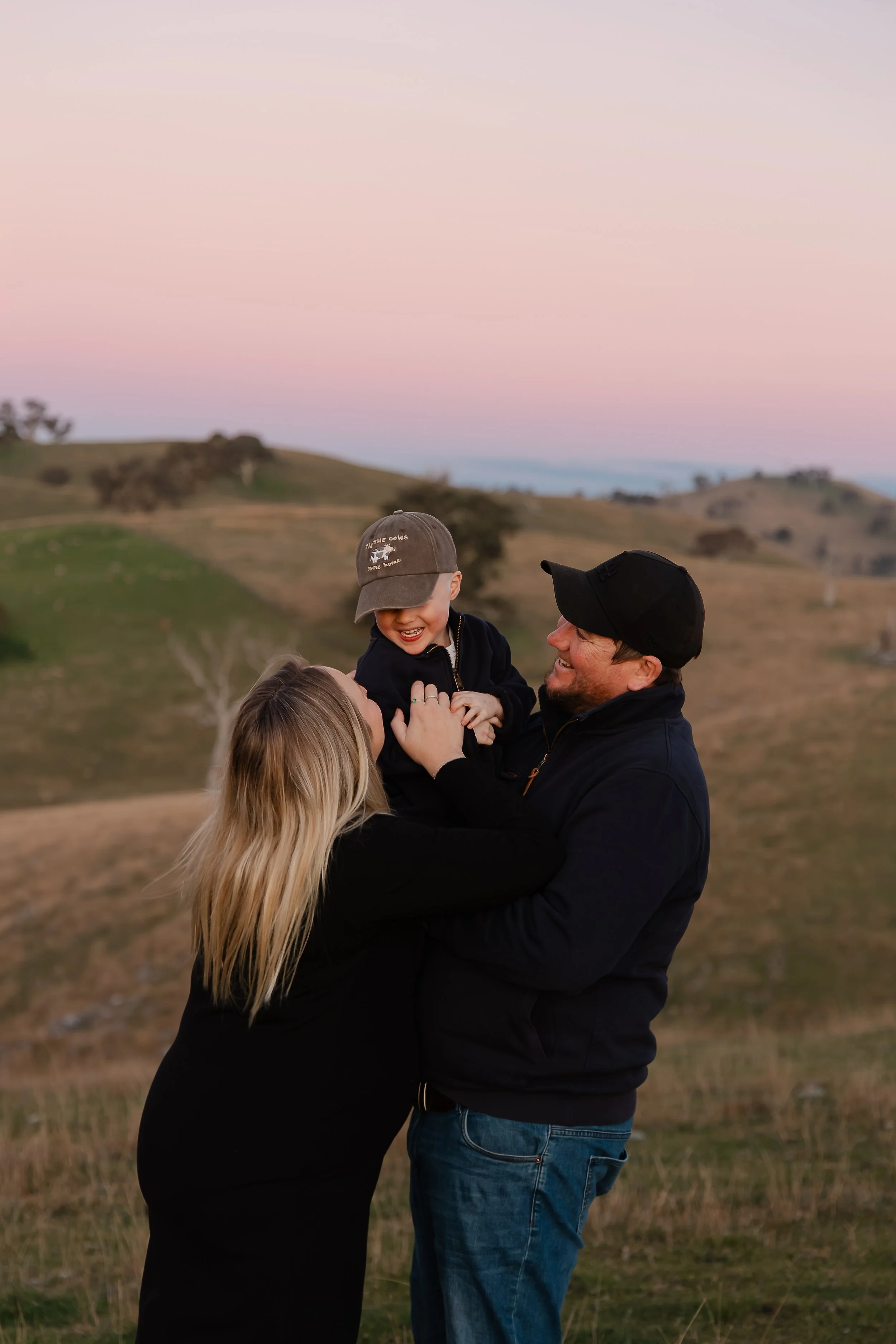 A happy family outdoors during sunset, with rolling hills and trees in the background. The man is holding a young child, and a woman with long blonde hair is embracing them.