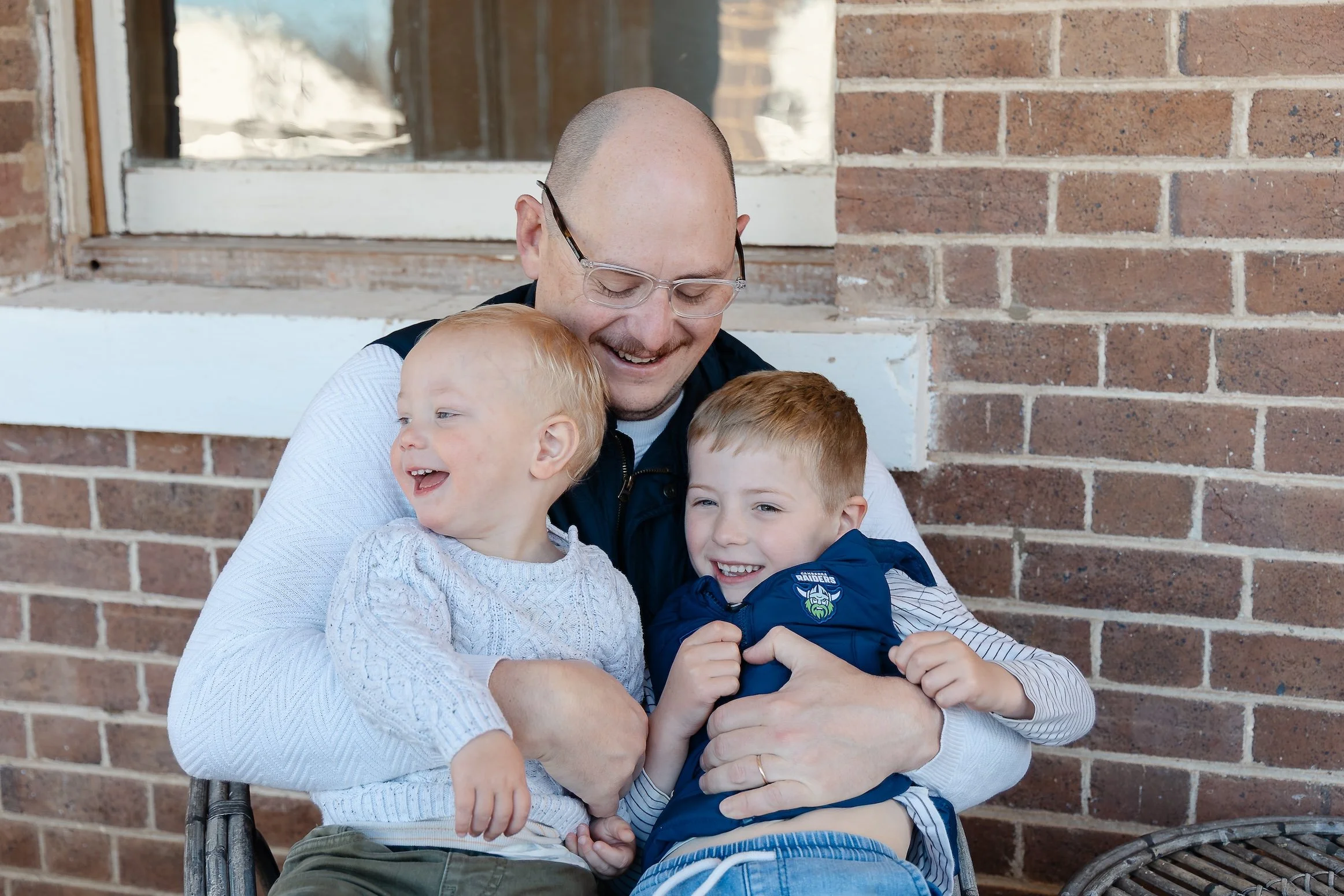 A man and three children hugging each other in front of a brick wall, smiling and enjoying a moment together on a sunny day.