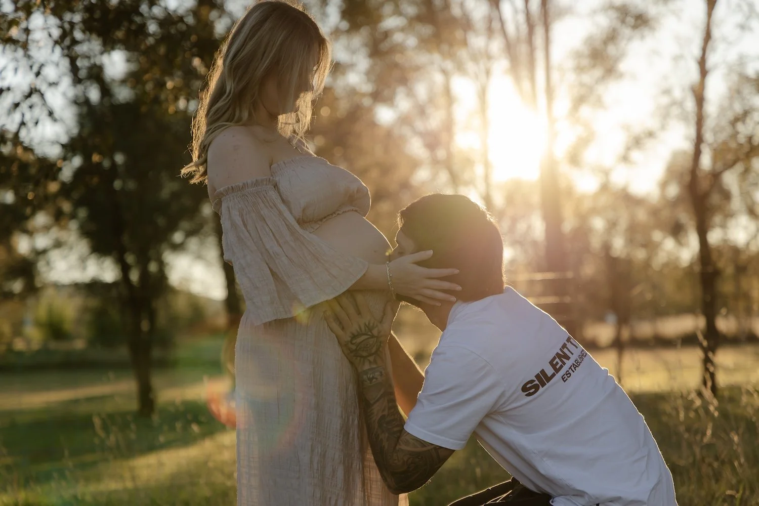 A pregnant woman with long blonde hair in a beige dress standing outdoors at sunset, holding her belly while a man with tattoos kneels before her, touching her face with both hands, in a romantic pose with trees and sunlight in the background.