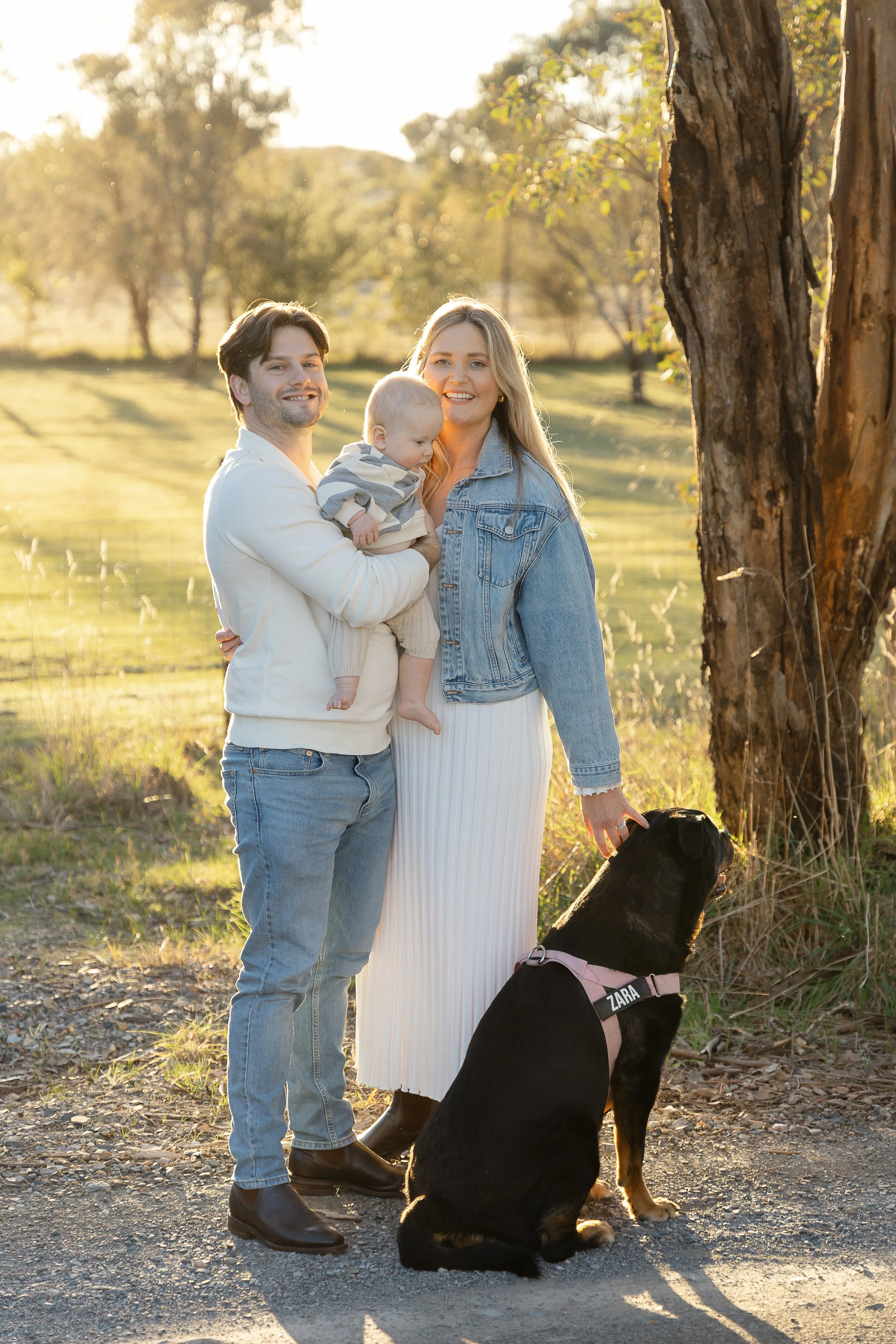 A family of three with a black service dog standing outdoors during golden hour. The man, woman, and baby are smiling; the woman is holding the baby, and the dog is sitting beside them wearing a harness labeled "ZARA." They are next to a tree in a gr