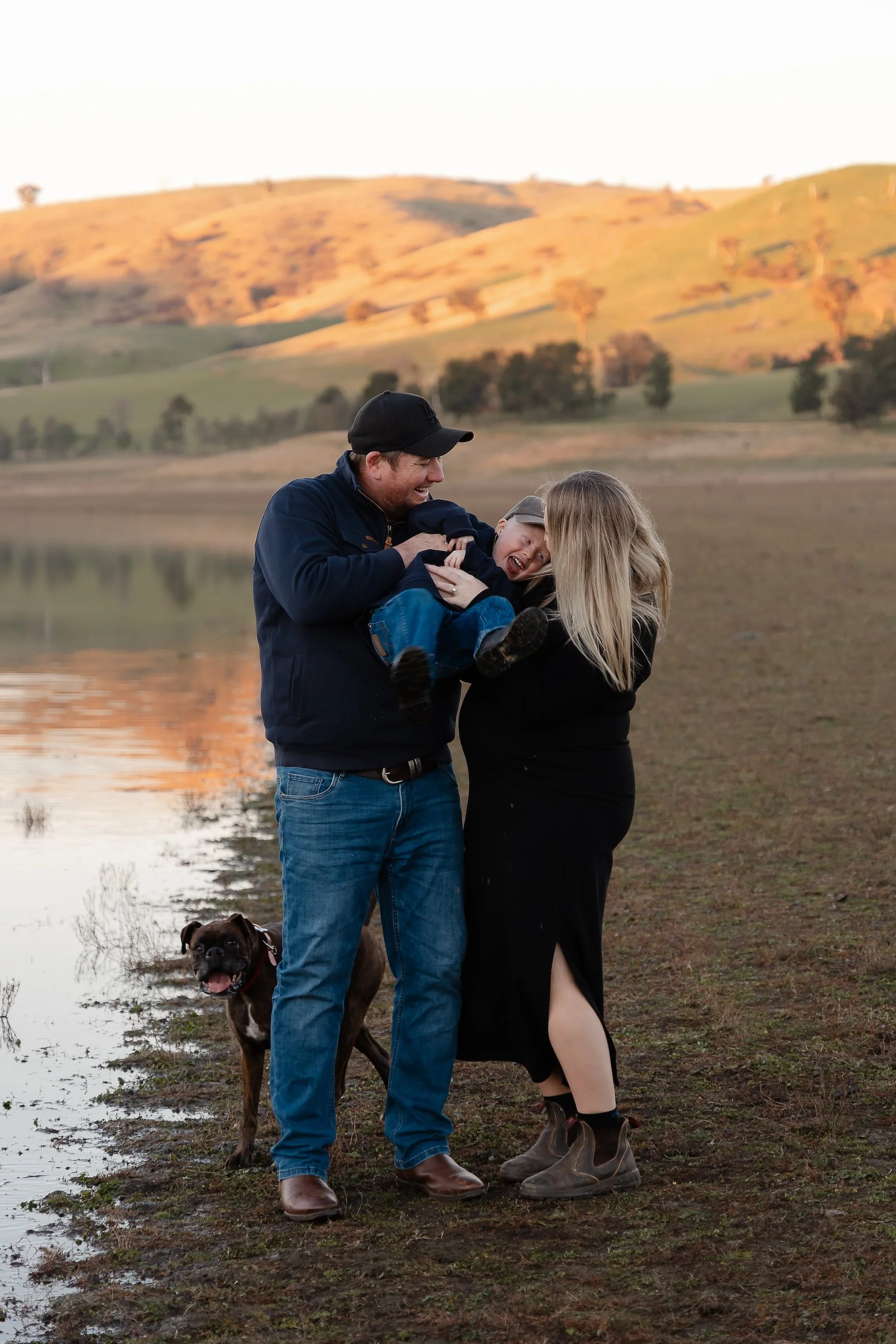 A family of three, a man, a woman, and a young boy, standing near a lake with a dog, in a rural landscape during sunset, with rolling hills in the background.